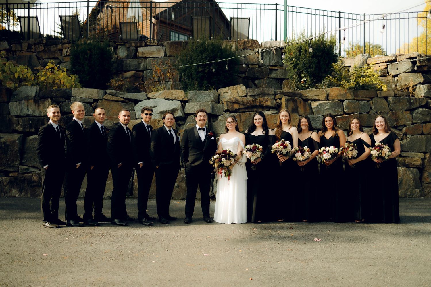 Wedding party in formal attire poses outdoors against stone wall backdrop at sunset.