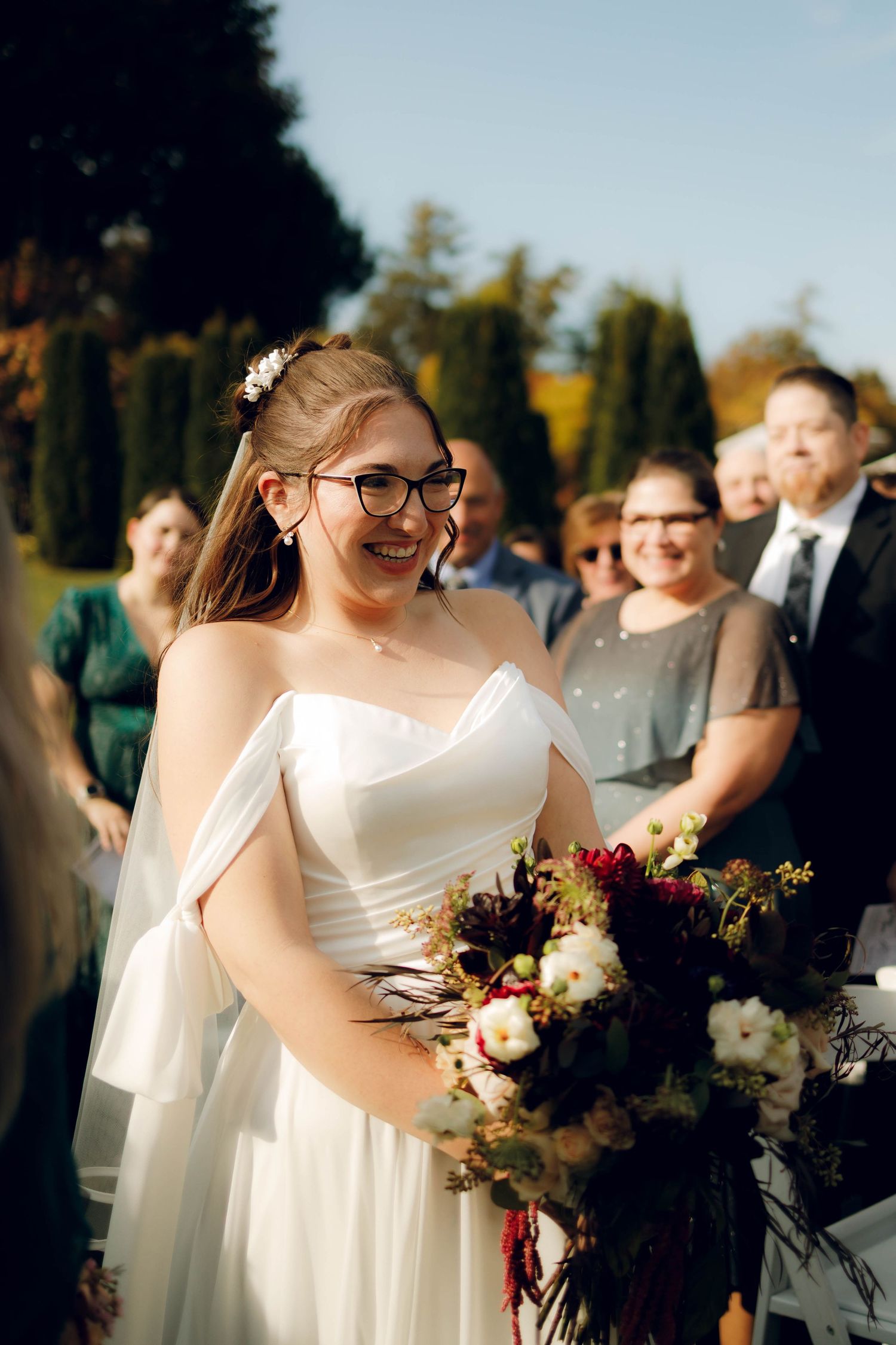 A bride in a white dress and glasses holds a beautiful bouquet of burgundy and white flowers at an outdoor wedding ceremony.