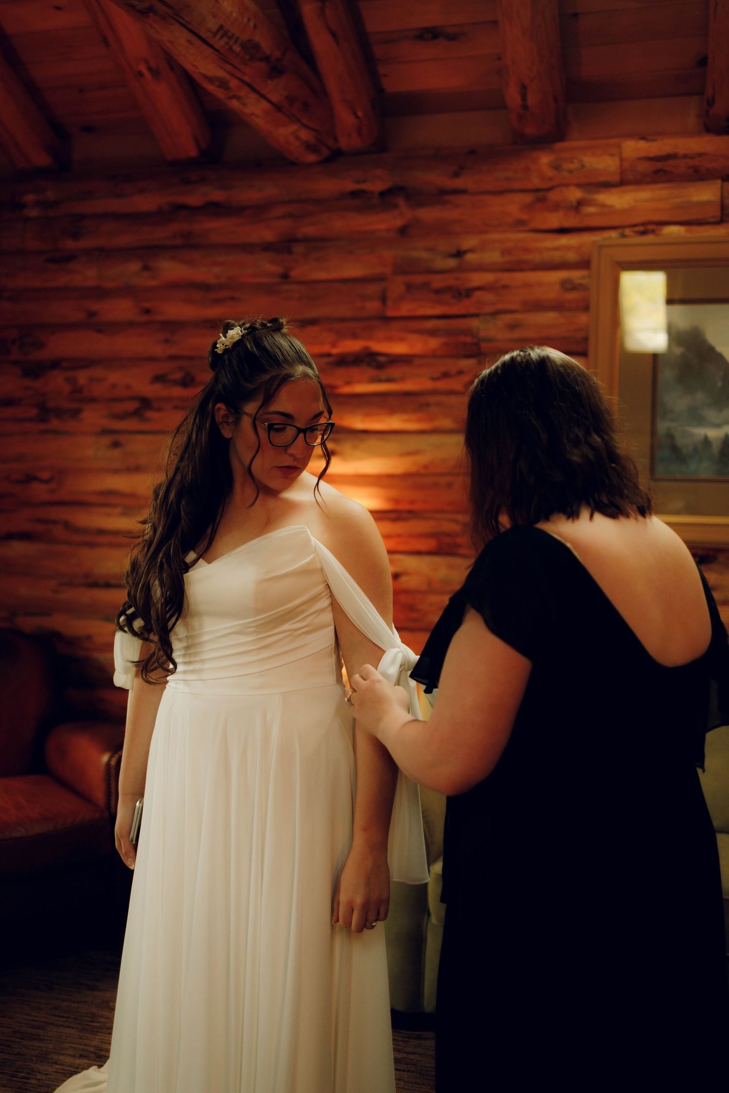 A person in a white wedding dress gets help with final adjustments inside a rustic log cabin setting.