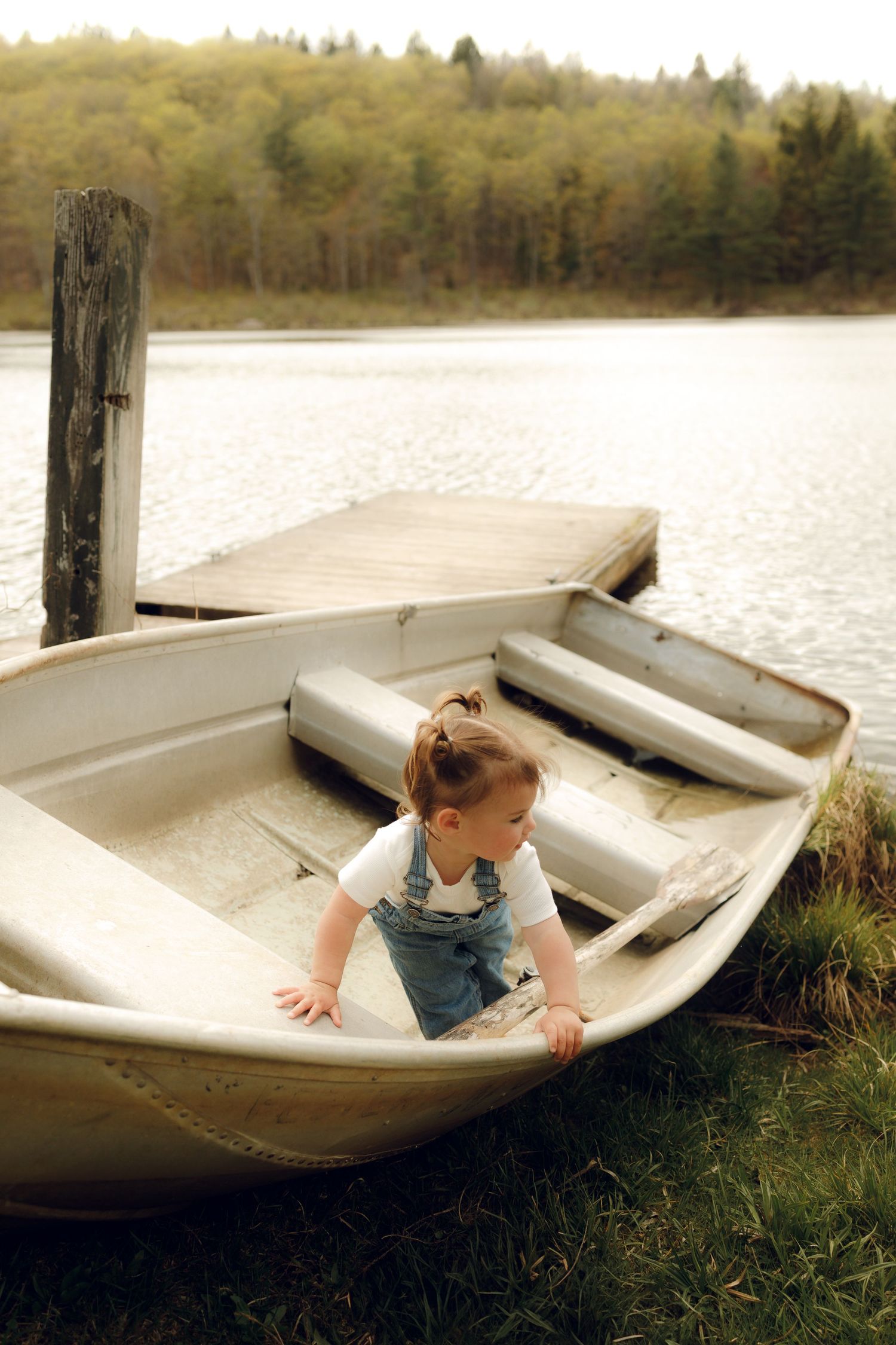 A small child wearing overalls explores a white rowboat docked by a peaceful lake on an autumn day.