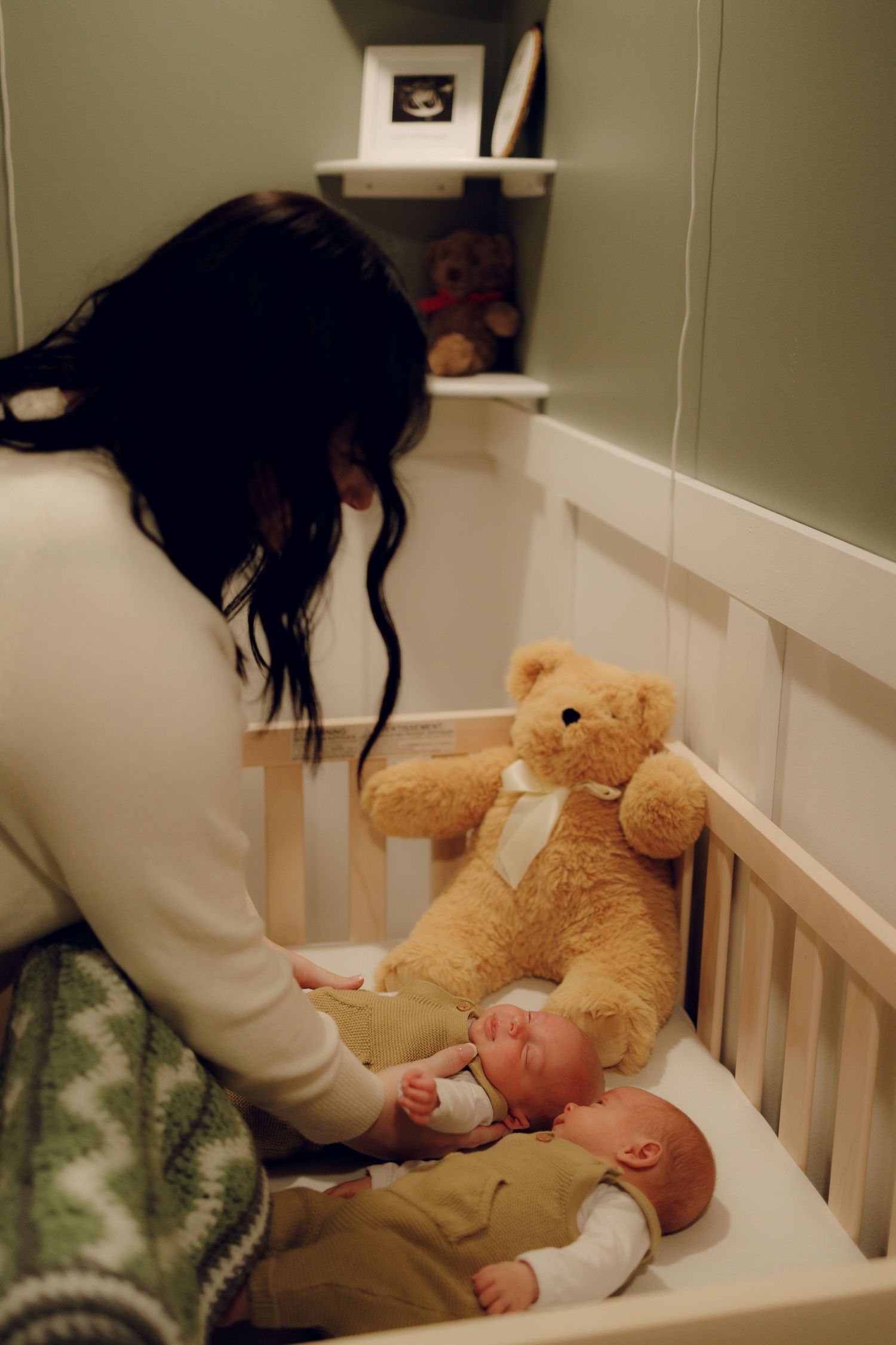 Parent comforting a small baby in white crib with teddy bear at bedtime.
