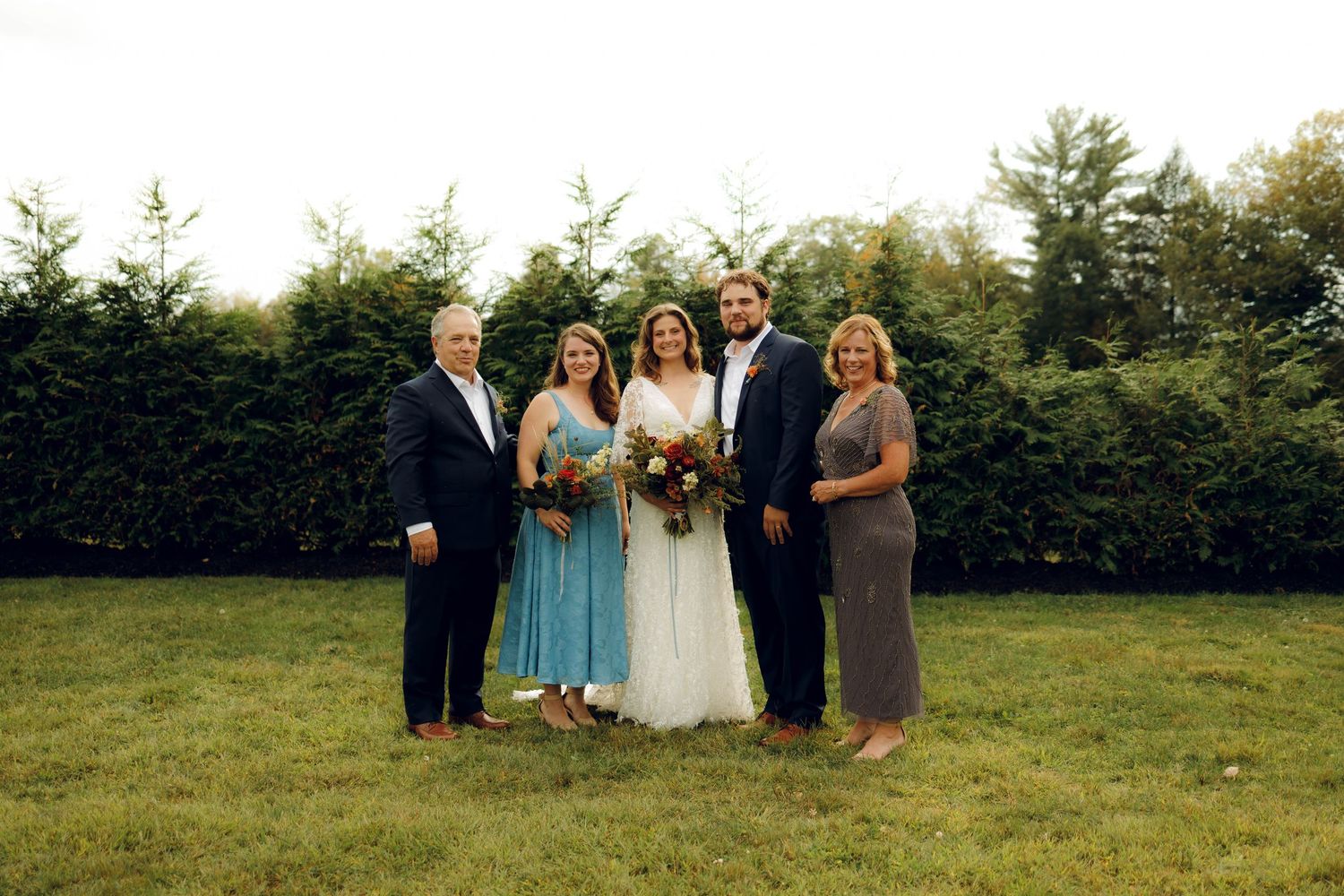 A wedding party poses together on a green lawn in front of a hedge on a sunny day.