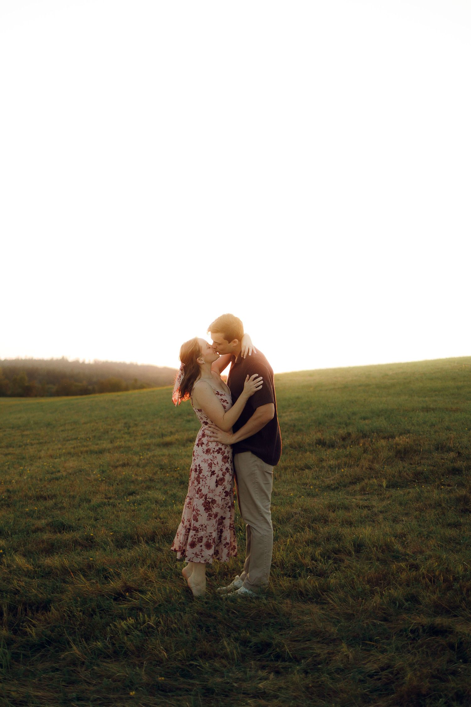 A couple embraces in a romantic sunset moment on a grassy hilltop as golden light bathes the pastoral landscape.