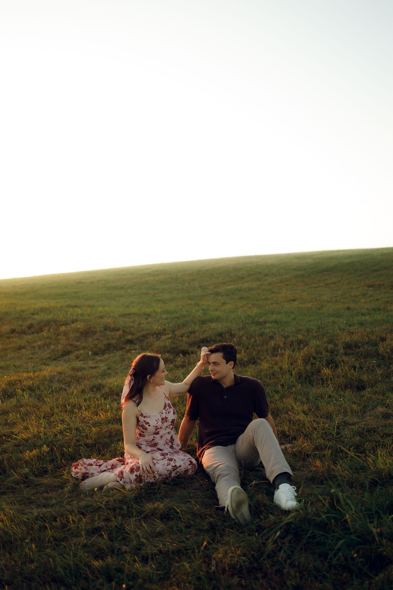 A couple relaxes together on a grassy hillside during golden hour in a romantic outdoor setting.