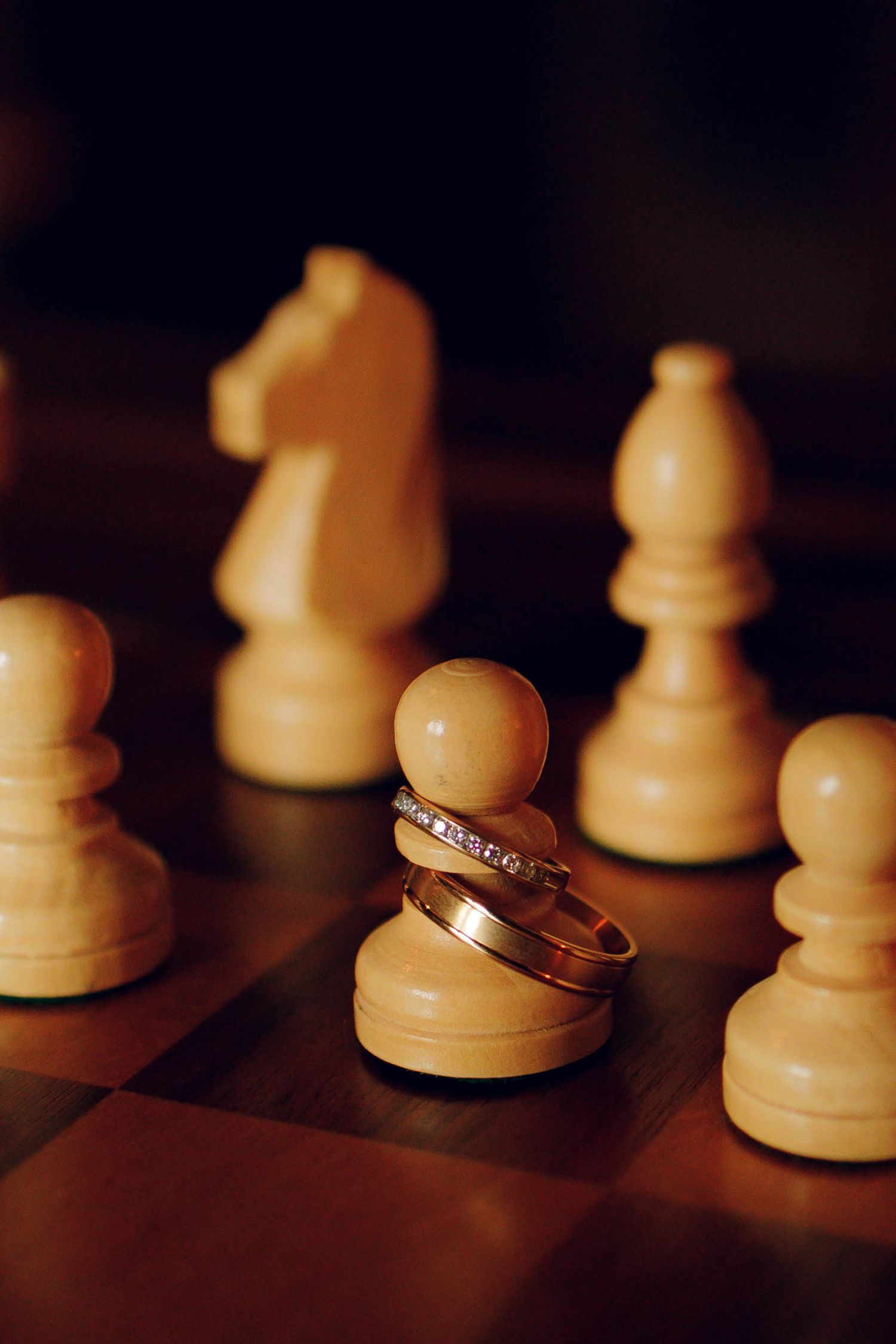 Wedding rings rest elegantly among white chess pieces on a dark wooden chessboard in dramatic lighting.