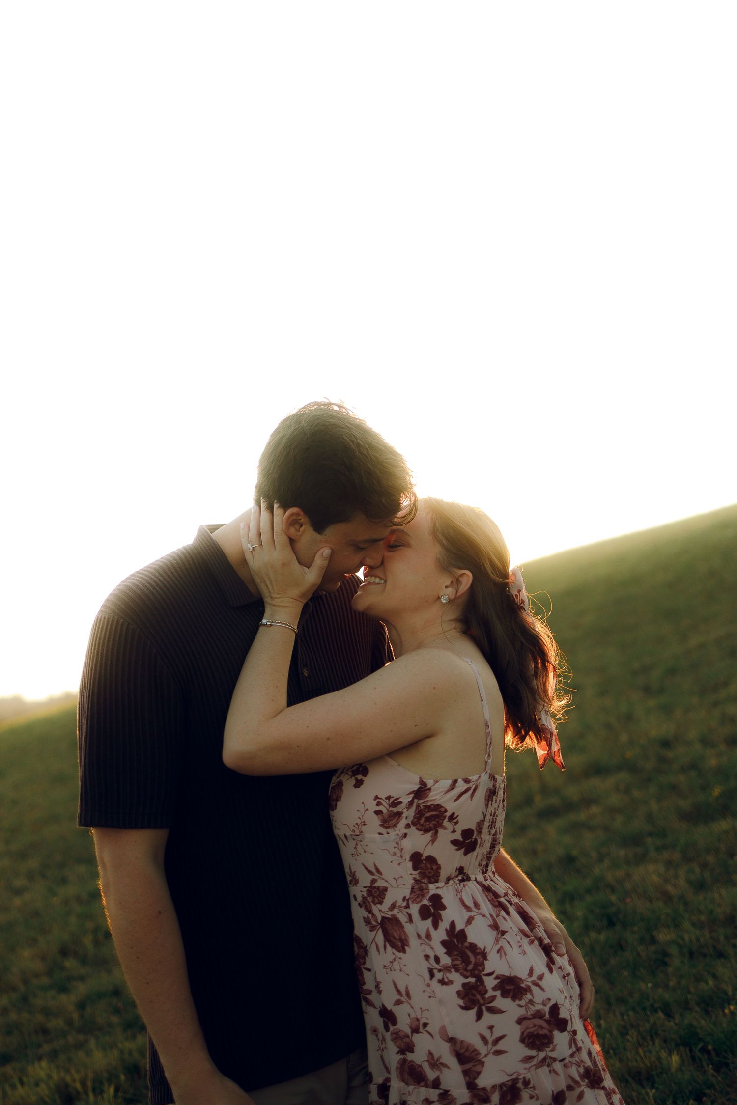 A couple shares a romantic kiss in a grassy field at sunset, creating a silhouetted scene against the golden sky.