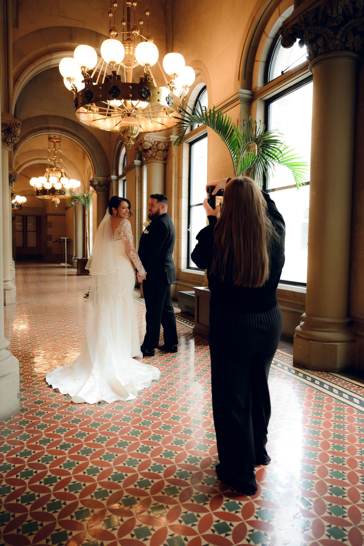 Panoramic view of an elegant ballroom with ornate chandeliers, arched windows, and patterned carpet showing silhouettes.