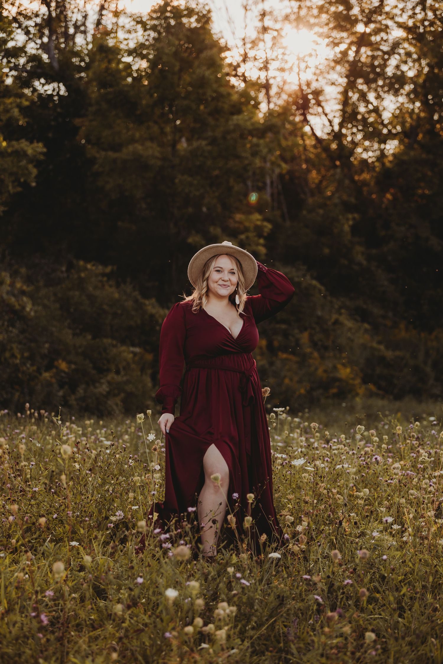 A figure in a burgundy dress sits gracefully among wildflowers in a meadow at golden hour.