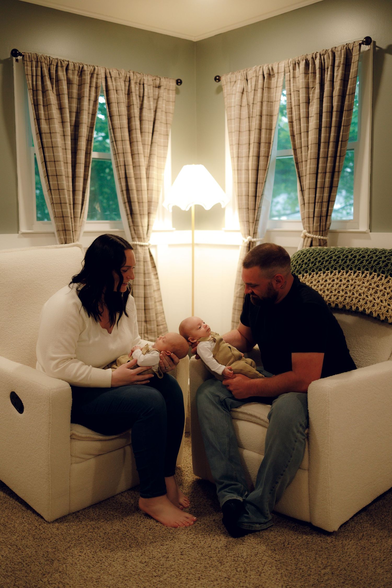 Two people sit together in white armchairs near a floor lamp, lovingly holding their baby in a cozy living room corner.