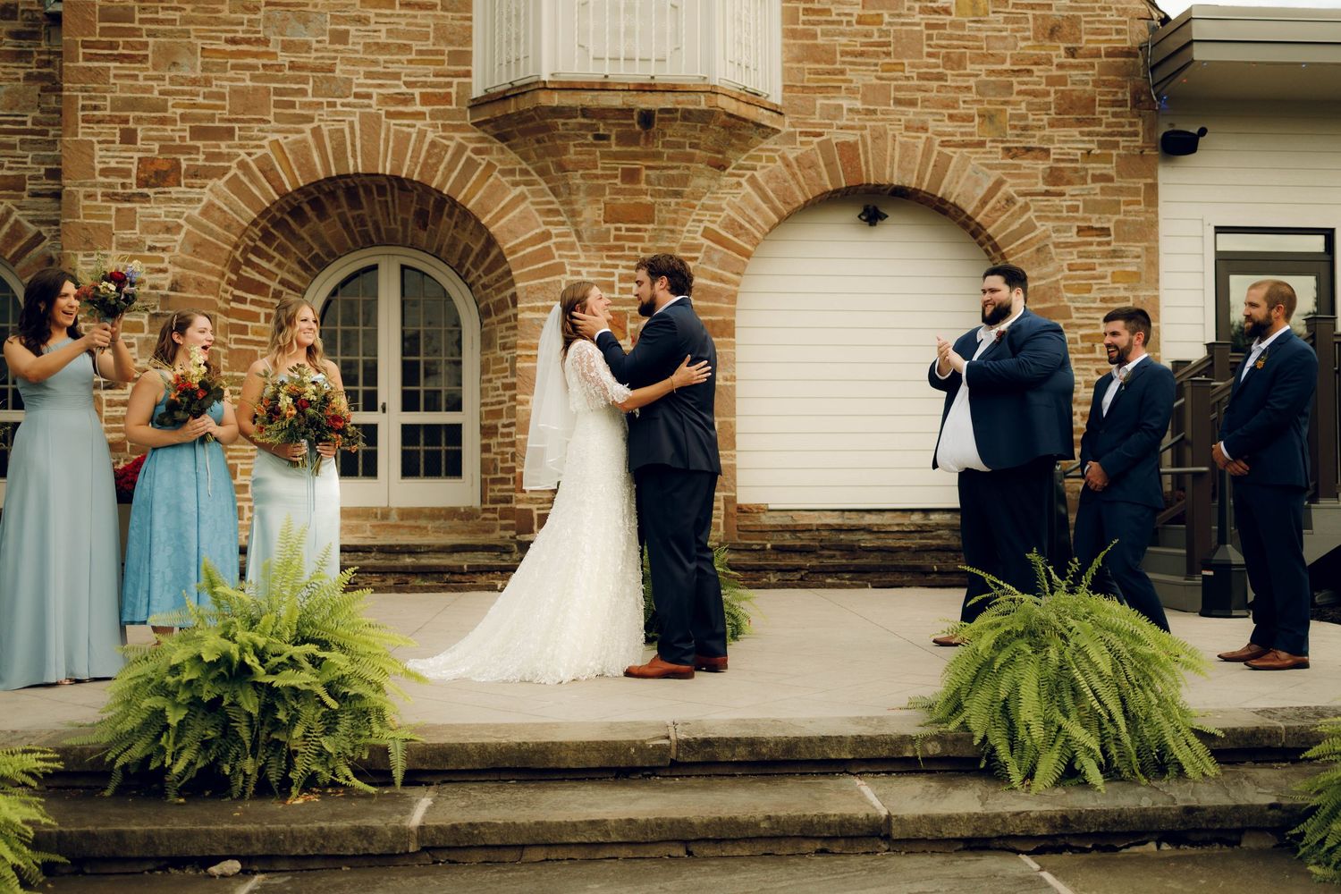 Elegant wedding couple and wedding party pose together on brick porch with lush green plants.