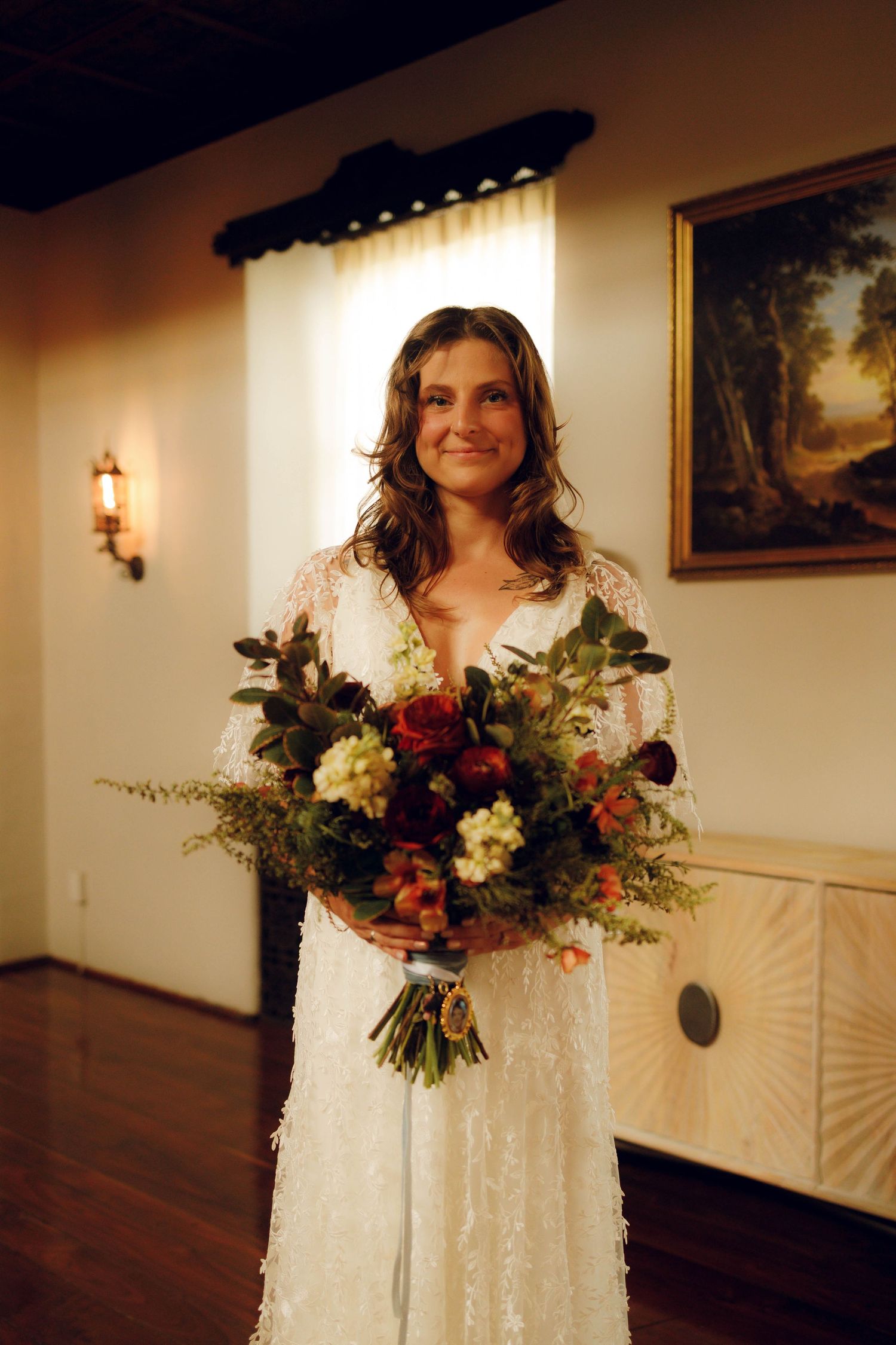 A bride in a white lace dress holds a vibrant autumn-colored bouquet with red and burgundy flowers in an indoor setting.