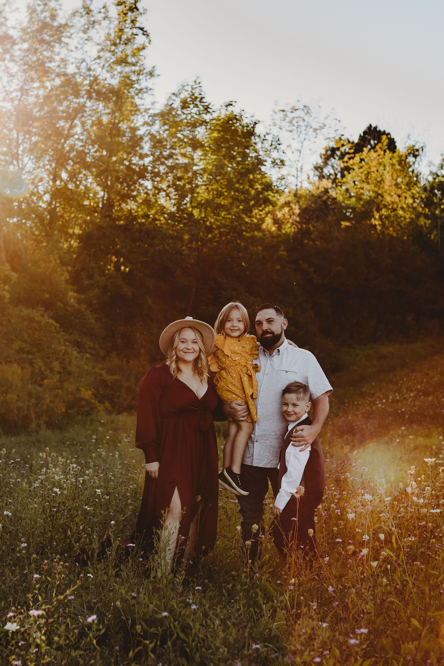 Family of four embracing in a sunlit meadow during golden hour as sunlight filters through autumn trees.