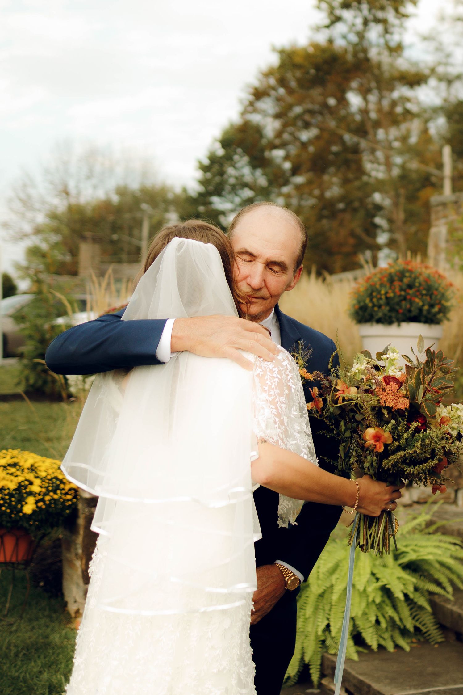 A couple in wedding attire embraces outdoors in autumn with colorful flowers and greenery in the background.