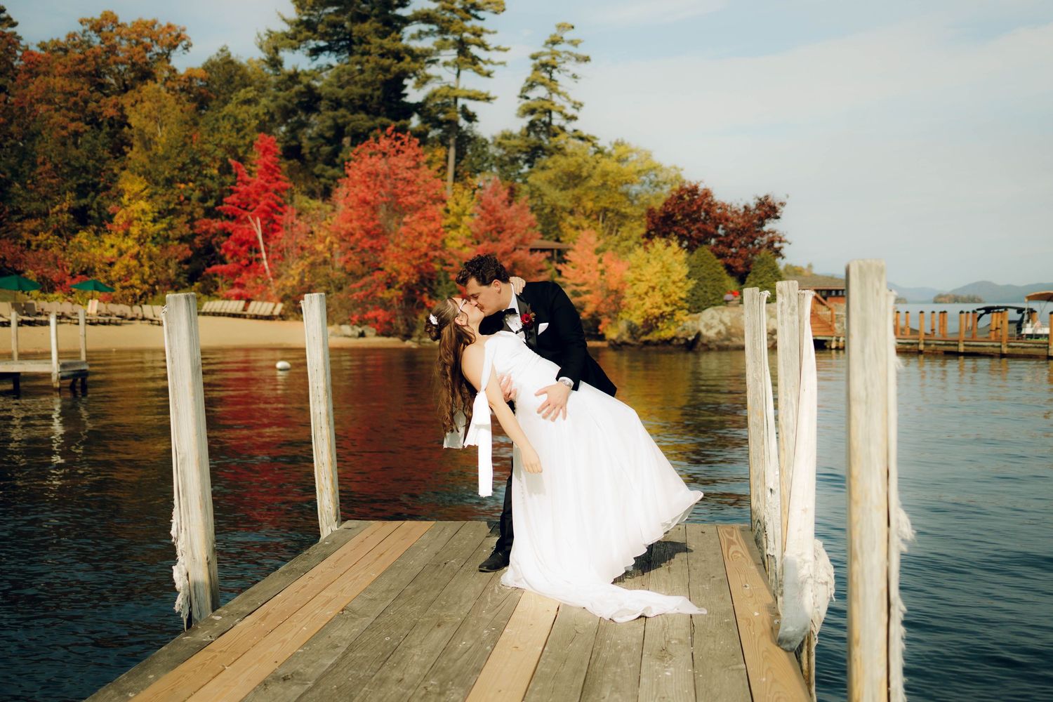 A couple in wedding attire sits on a wooden dock surrounded by autumn foliage and lake views.