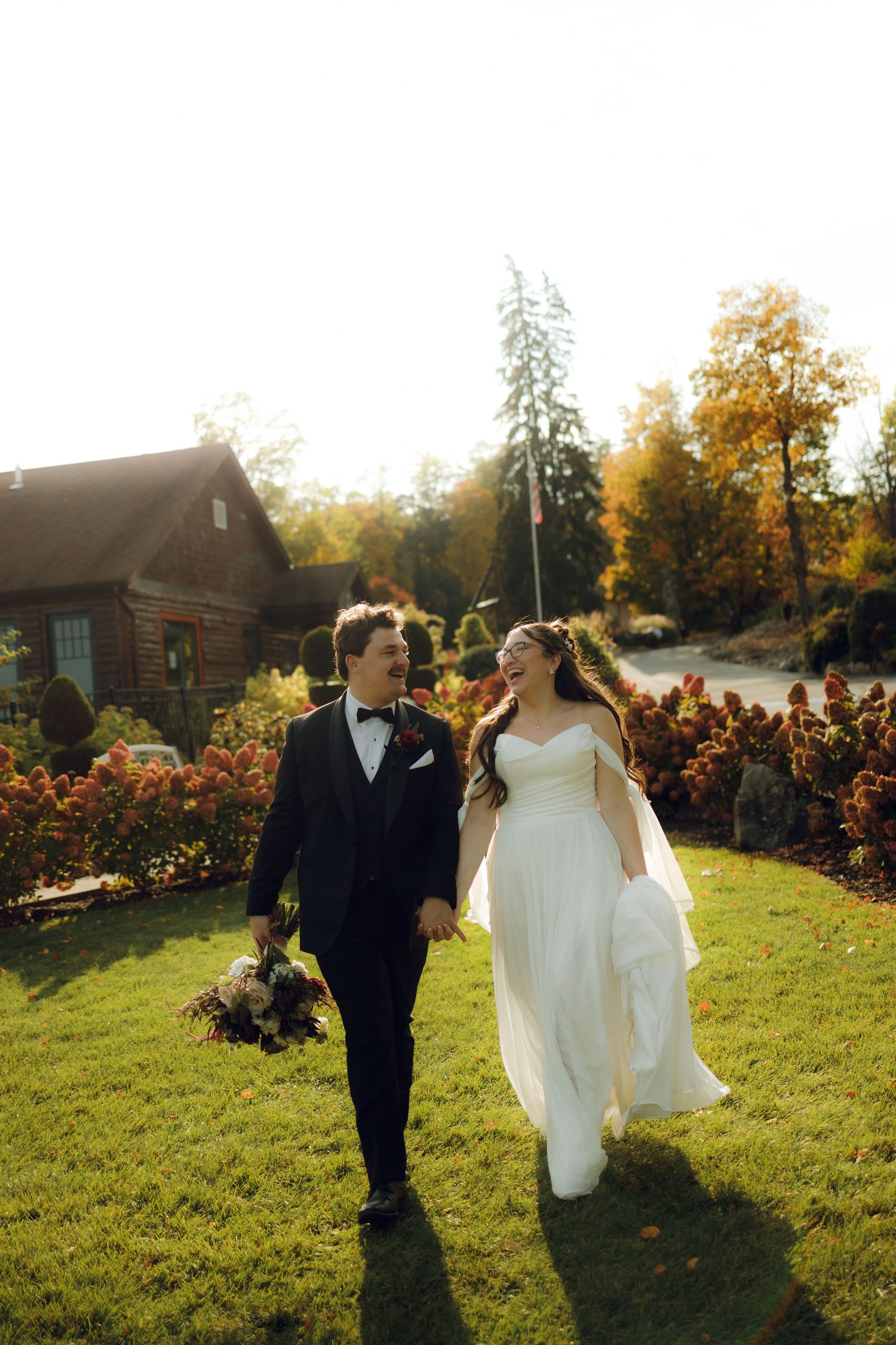 Newlywed couple walks through a sunlit garden with autumn trees and blooming flowers during golden hour.