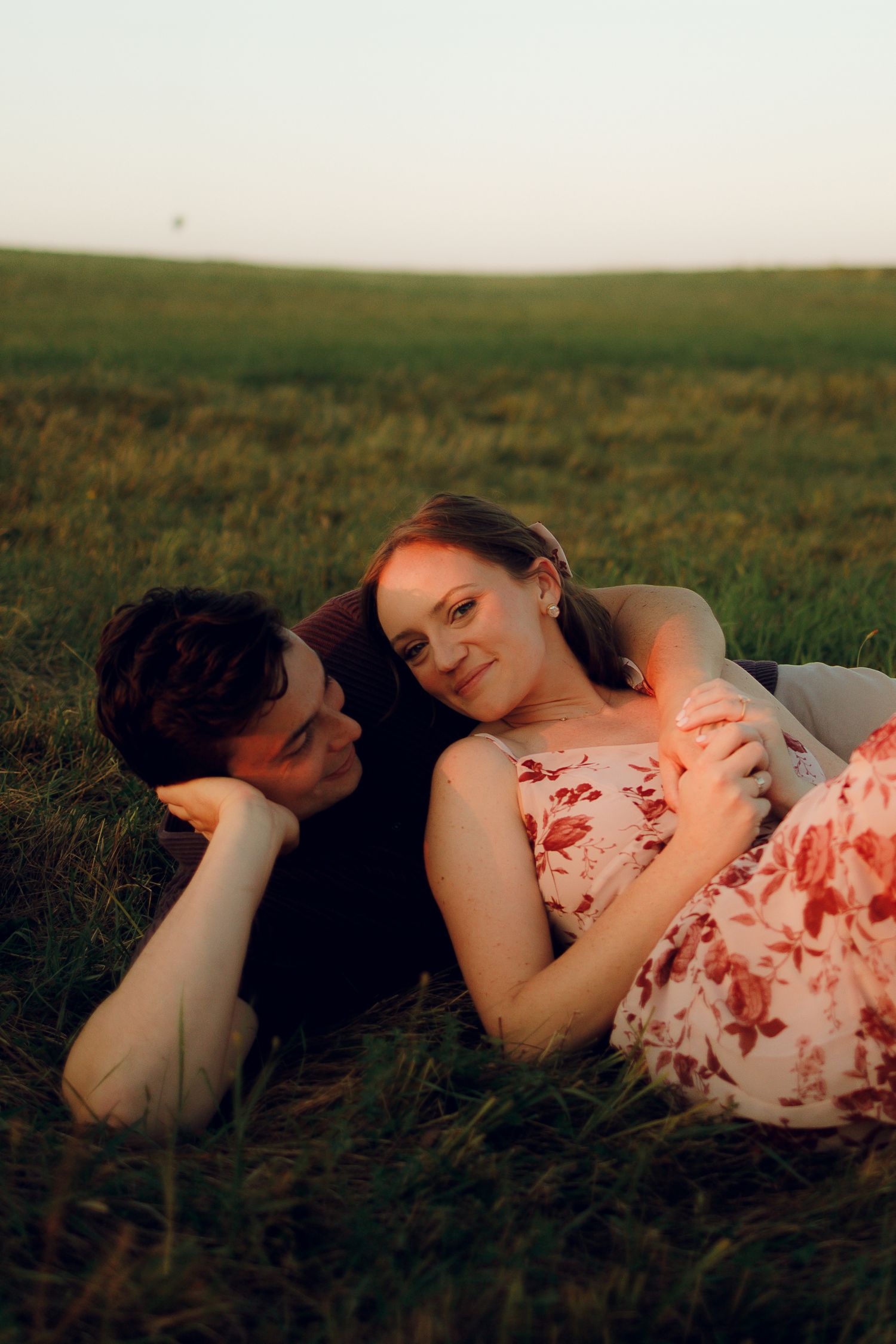A couple lies together in a grassy meadow at dusk wearing white and floral clothing.