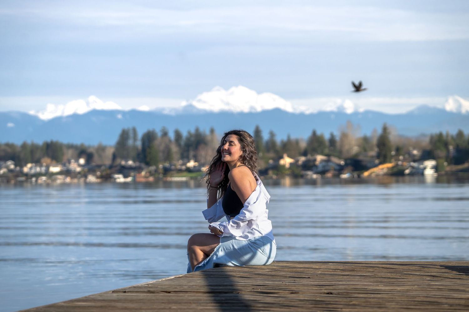 Person enjoying a moment on lakeside dock with mountains and flying bird in background.