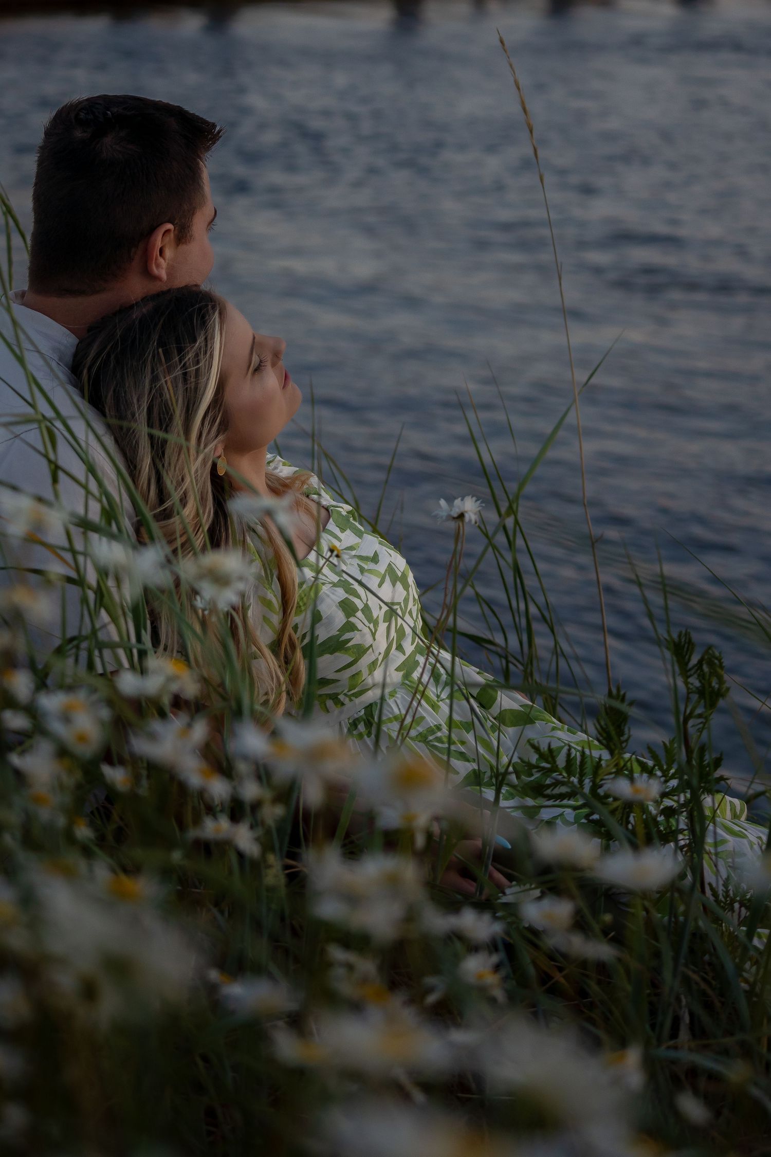 A couple embraces among waterside daisies at dusk with a marina in the background.