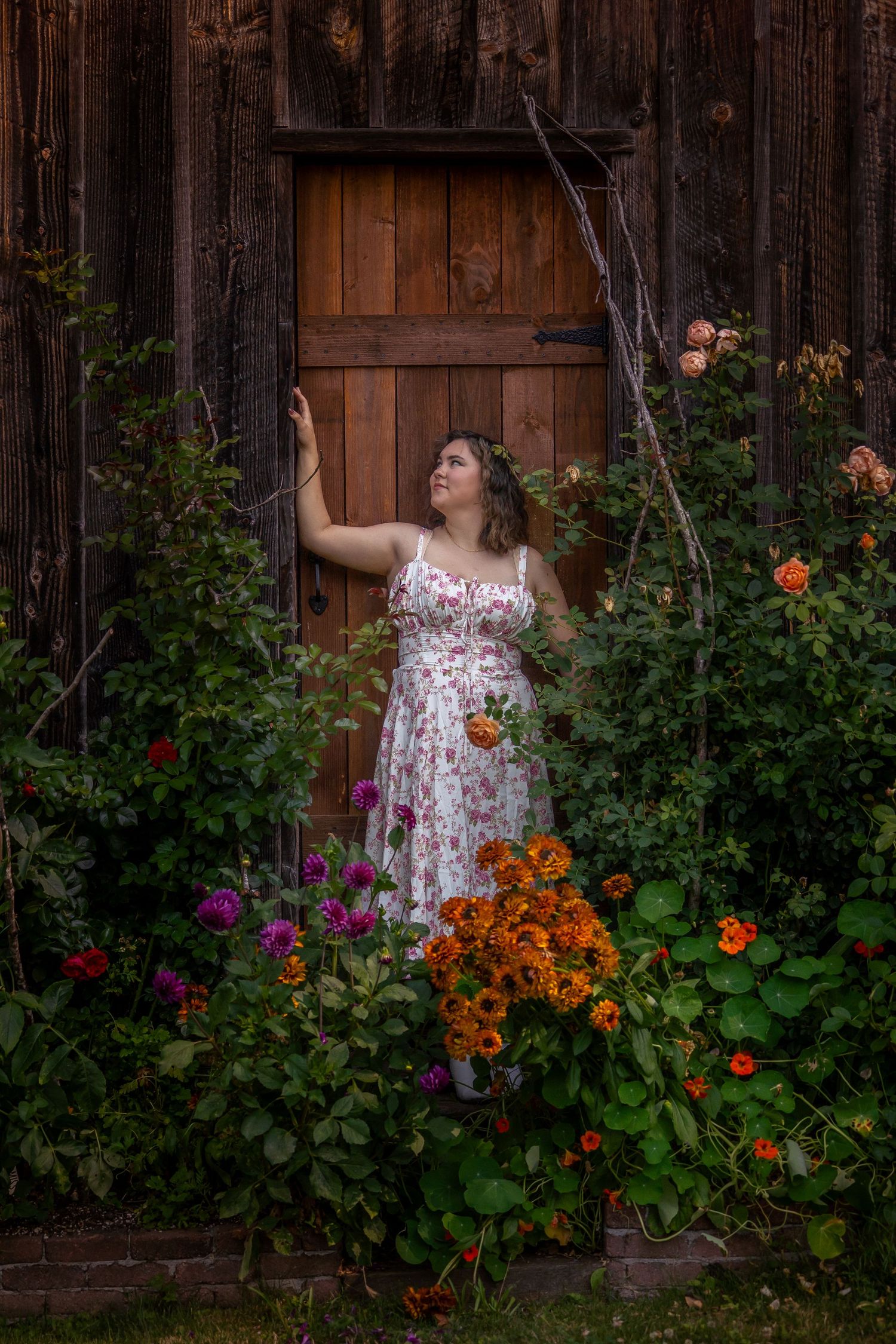 Person in floral dress posing by rustic wooden barn wall surrounded by colorful garden flowers.