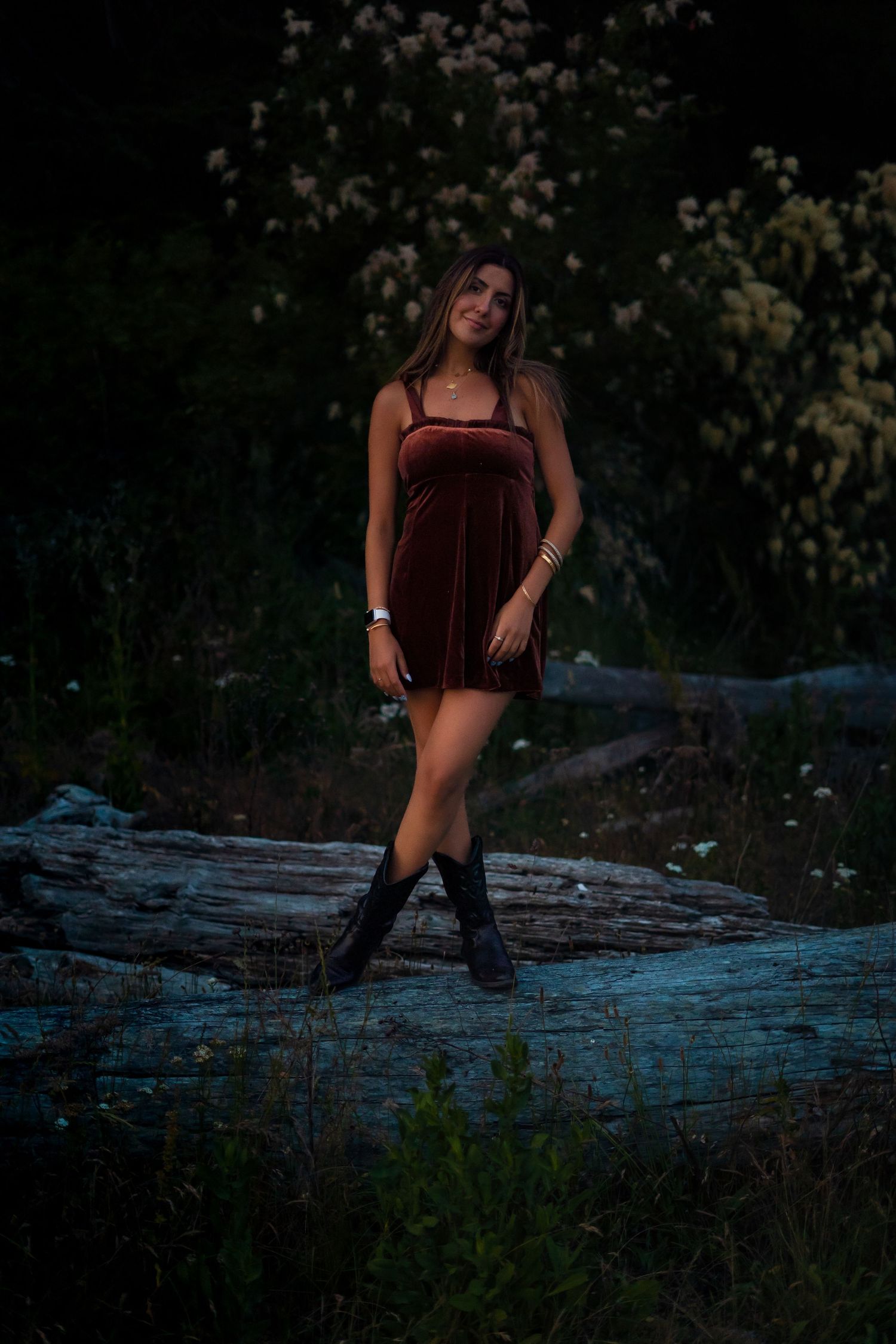 Person in velvet dress posing on fallen log at twilight among blooming flowers.