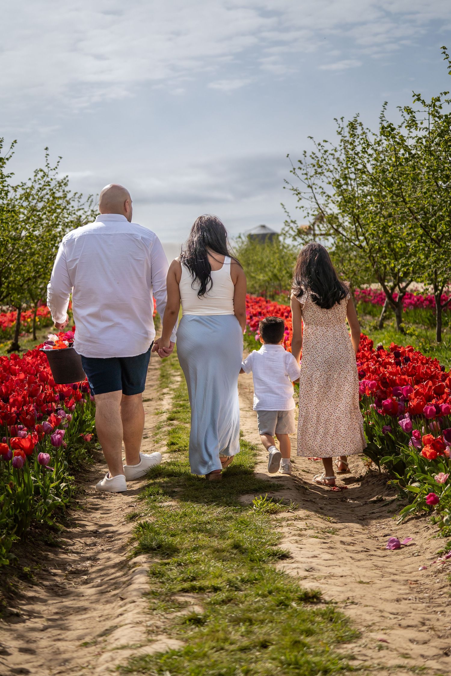 Family walking through vibrant red tulip field with trees and cloudy sky.