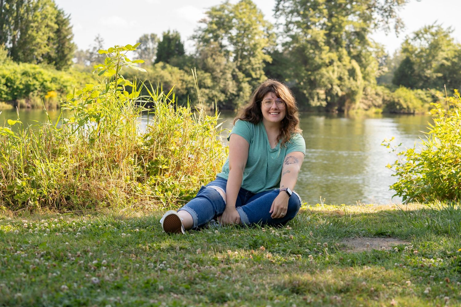 Person sitting on grass by a peaceful pond surrounded by lush summer greenery.