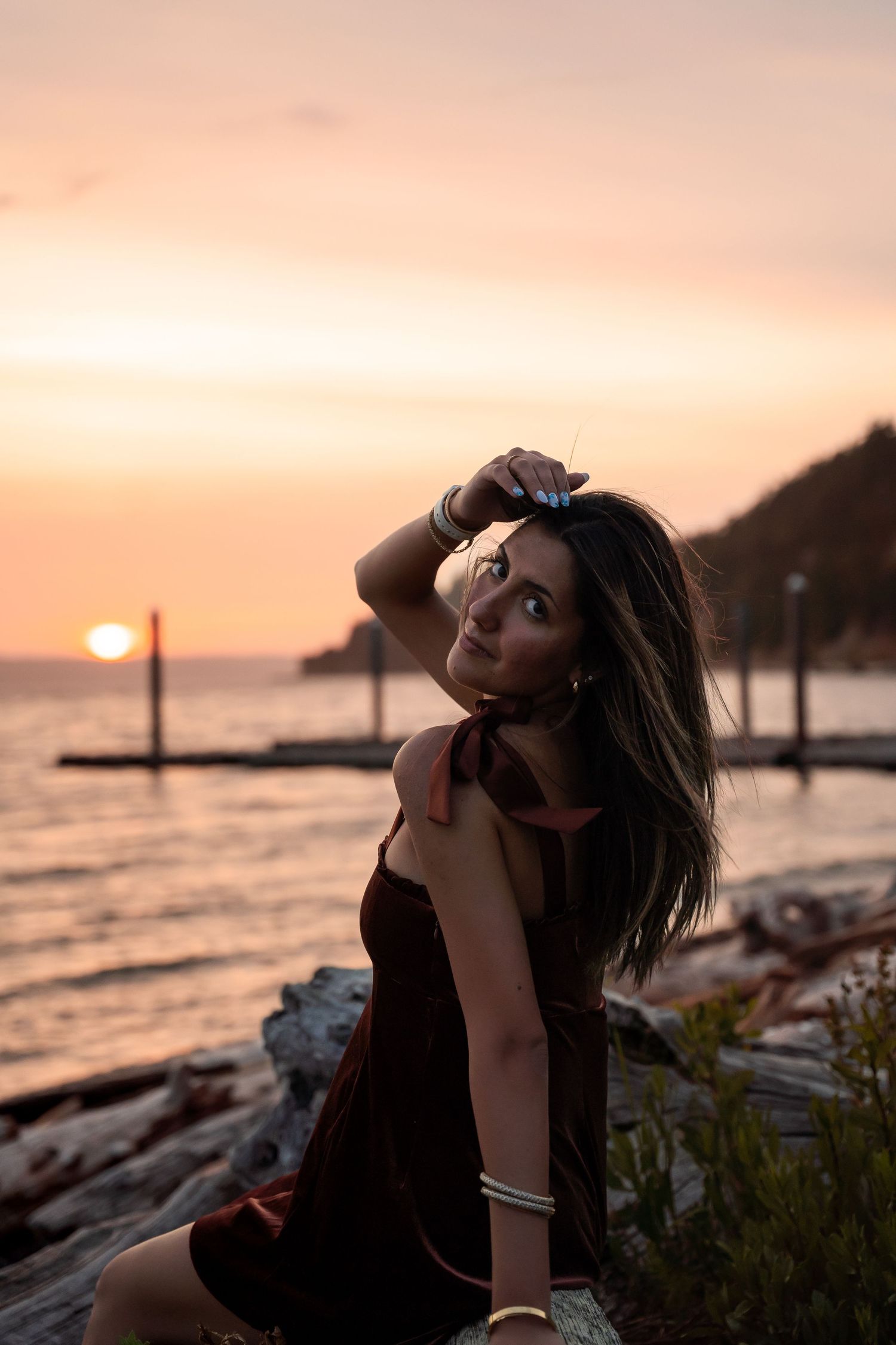 A silhouette sits by the ocean at sunset with a warm orange sky and pier in the background.