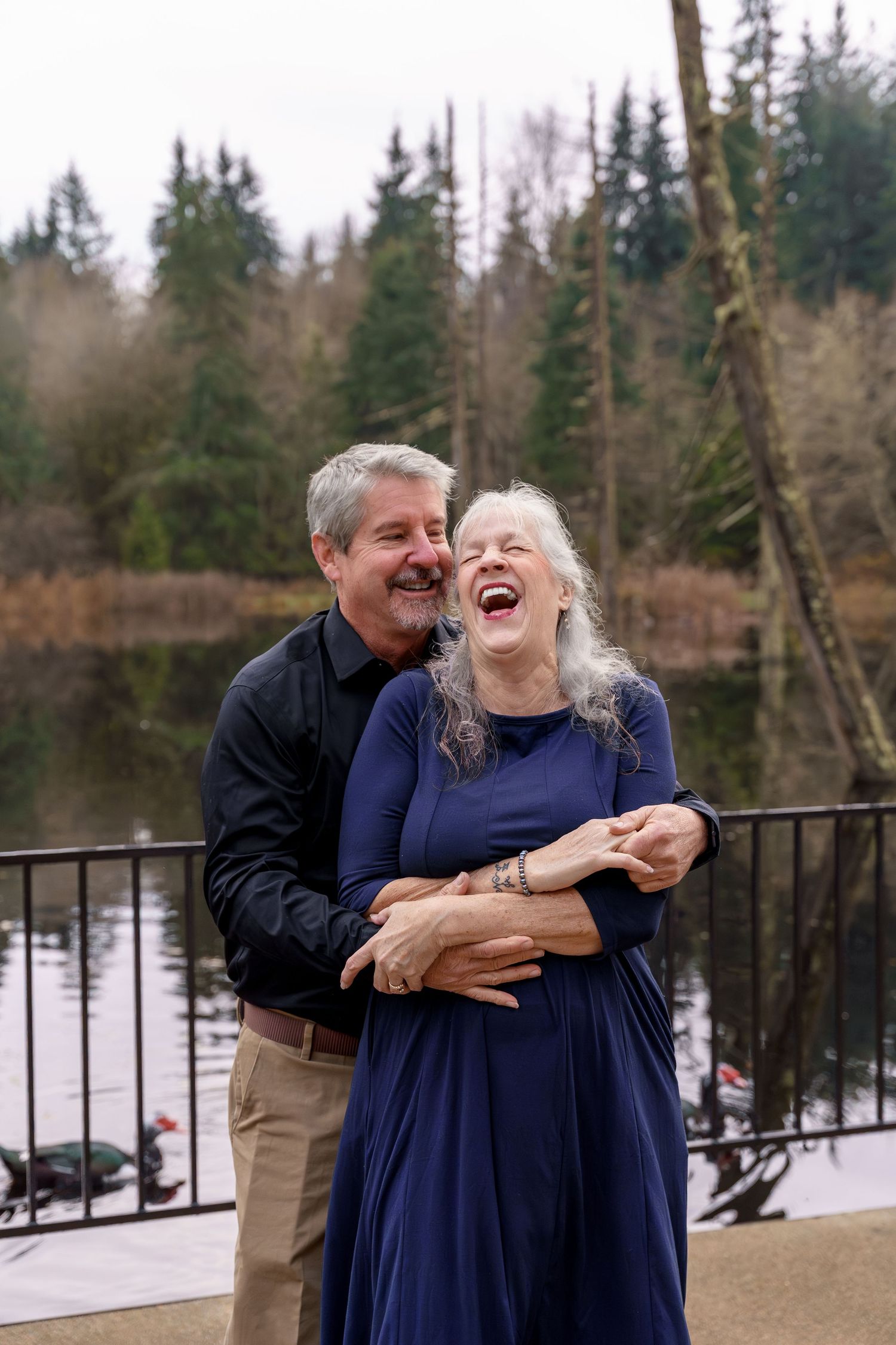A couple in navy blue outfits share a joyful embrace and laugh together on a deck overlooking a winter lake.