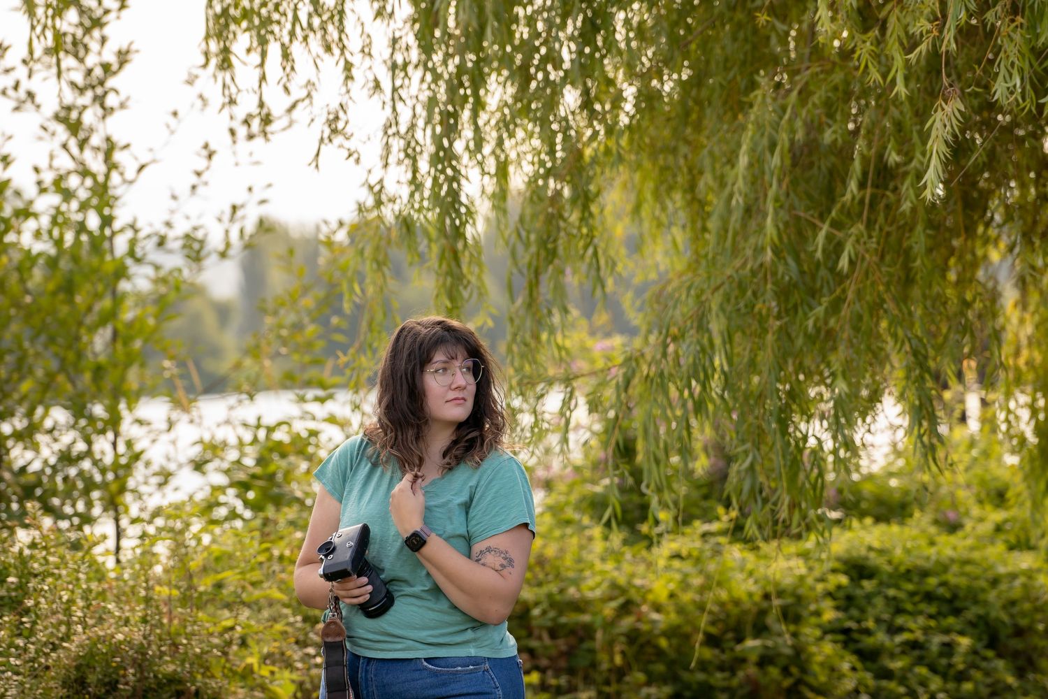 A photographer with a camera stands beneath drooping willow branches near a waterfront.