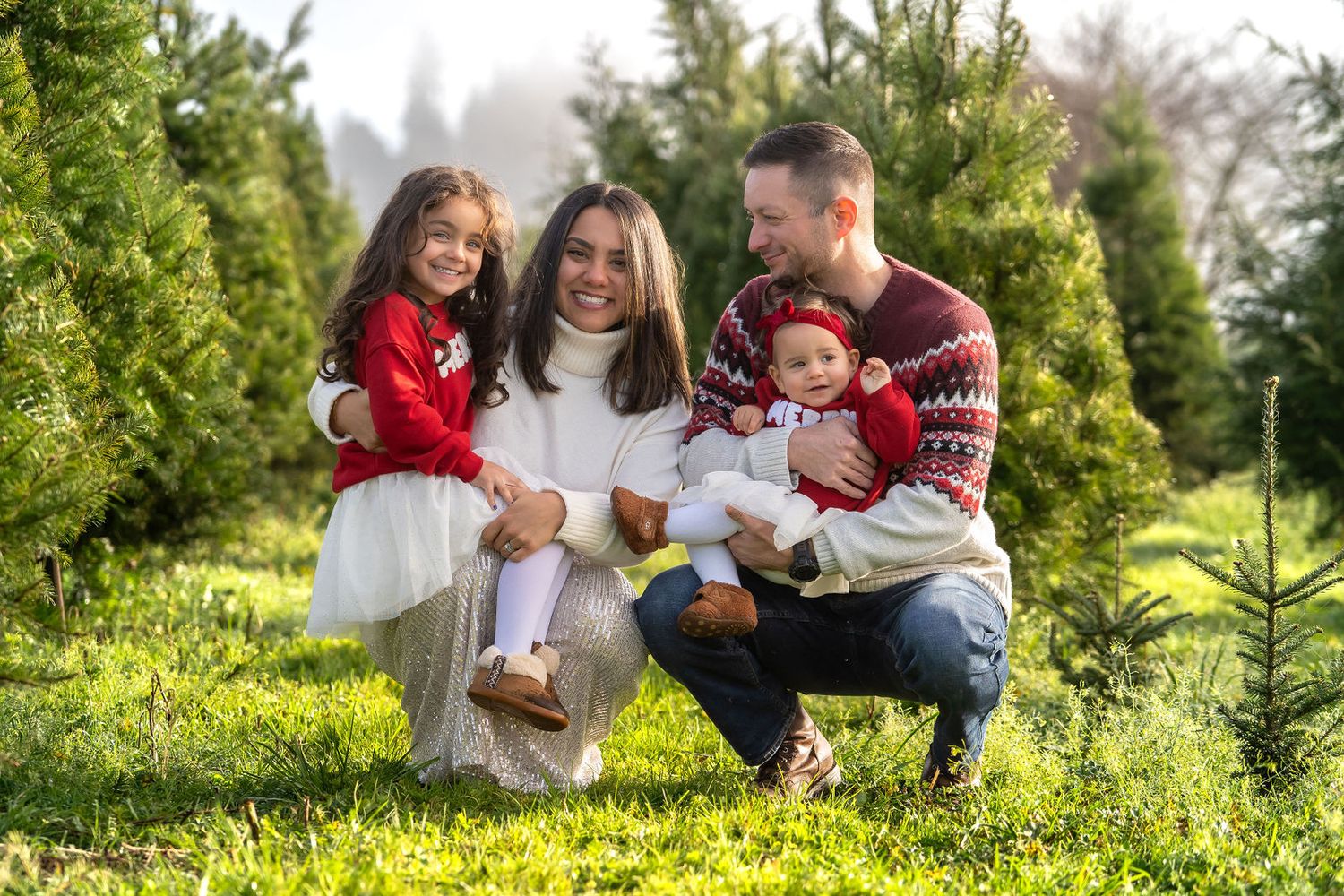 Family wearing festive red and white sweaters pose together among green Christmas trees on a sunny day.