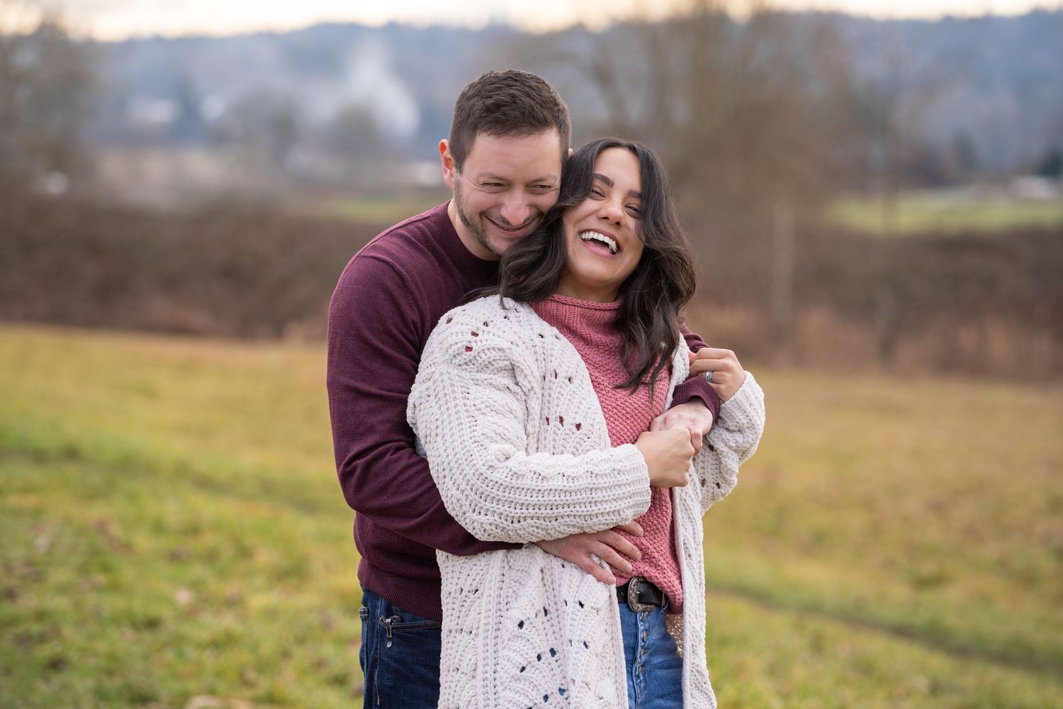 A couple shares a joyful candid moment together in a sweater and cardigan against an autumn field background.