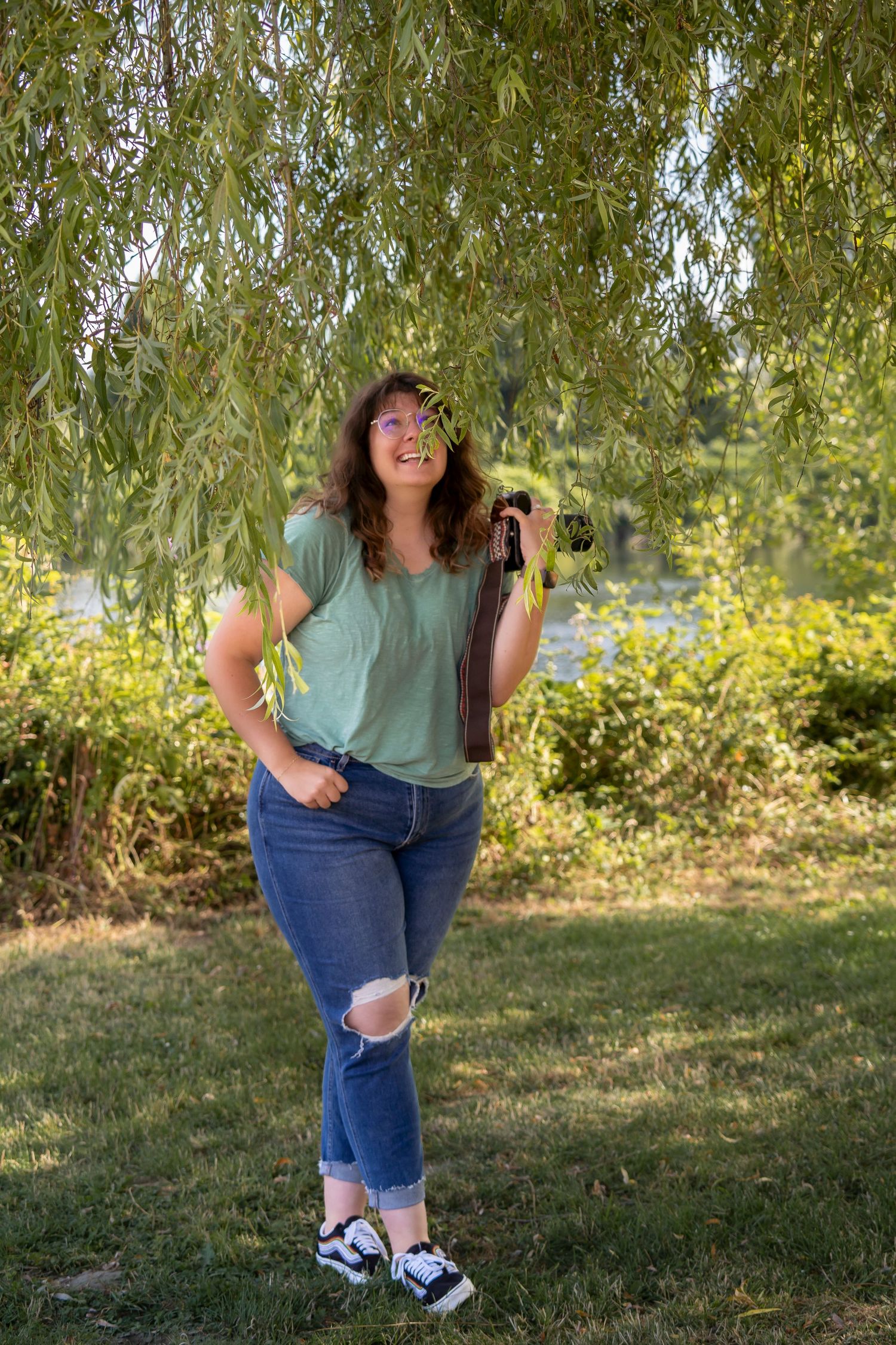 A photographer in casual attire smiles while holding a camera under a willow tree.