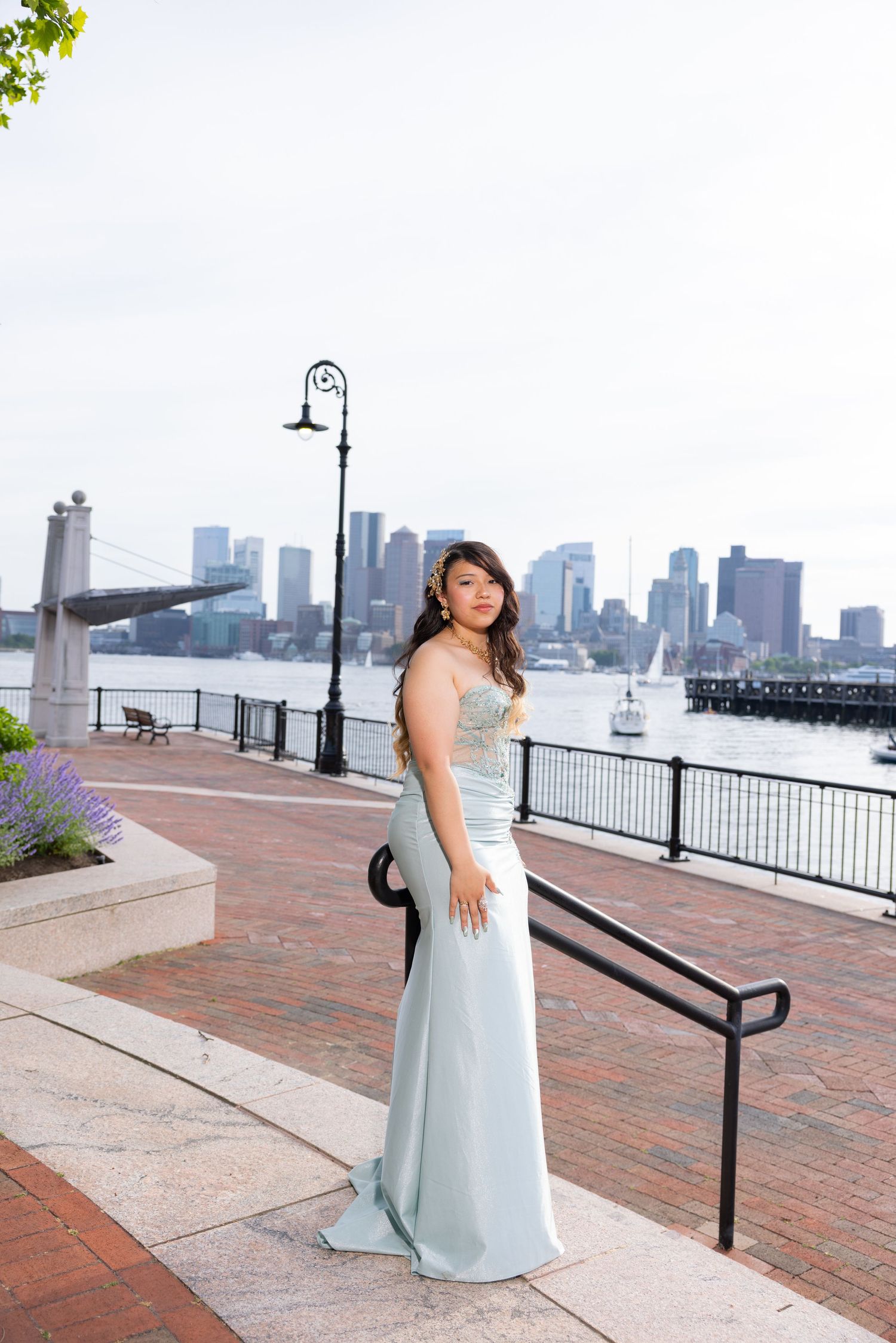Young woman in elegant light blue gown poses on brick waterfront promenade with Boston skyline in background.
