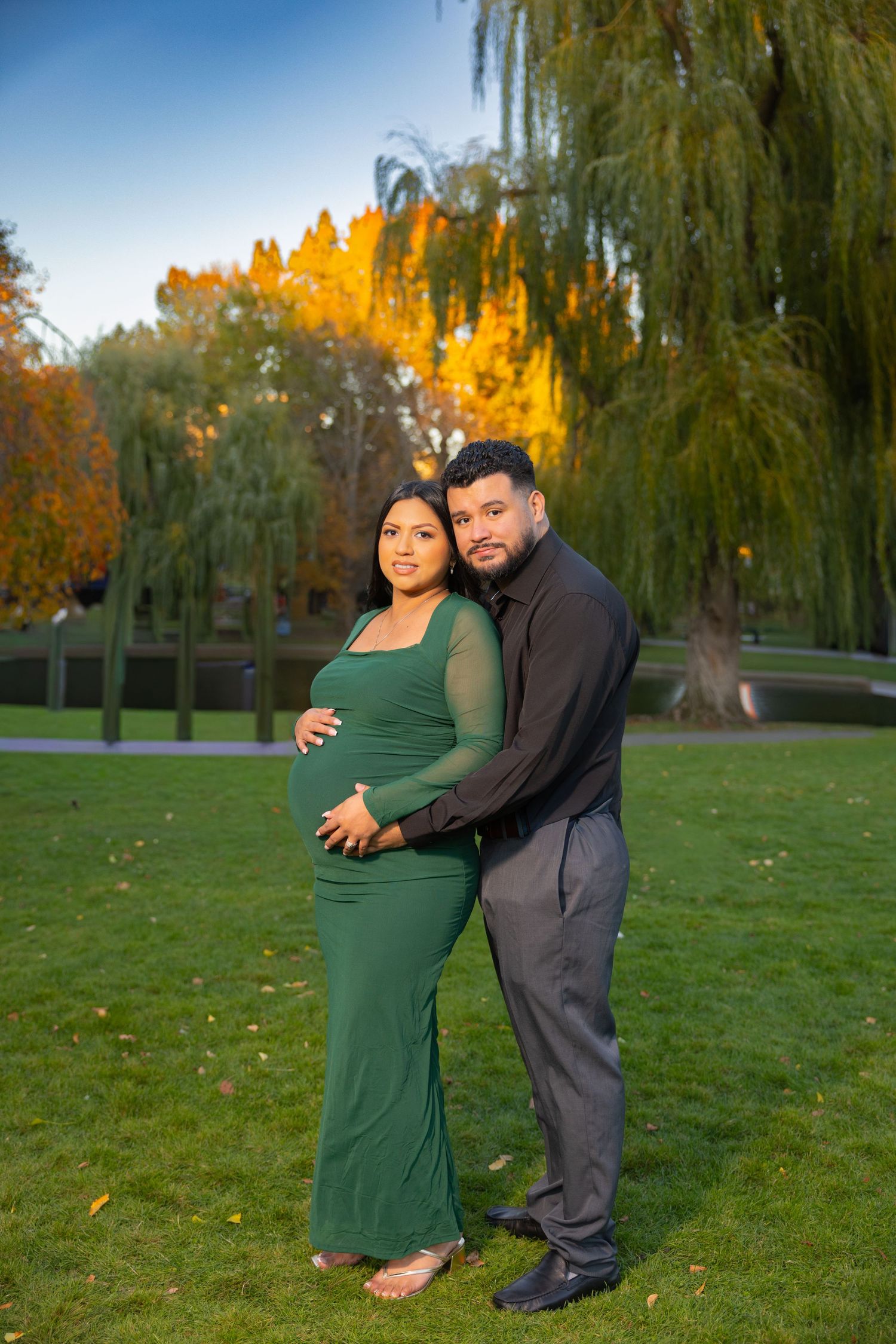 A couple embraces on a grassy lawn with autumn trees and weeping willows creating a romantic fall backdrop.