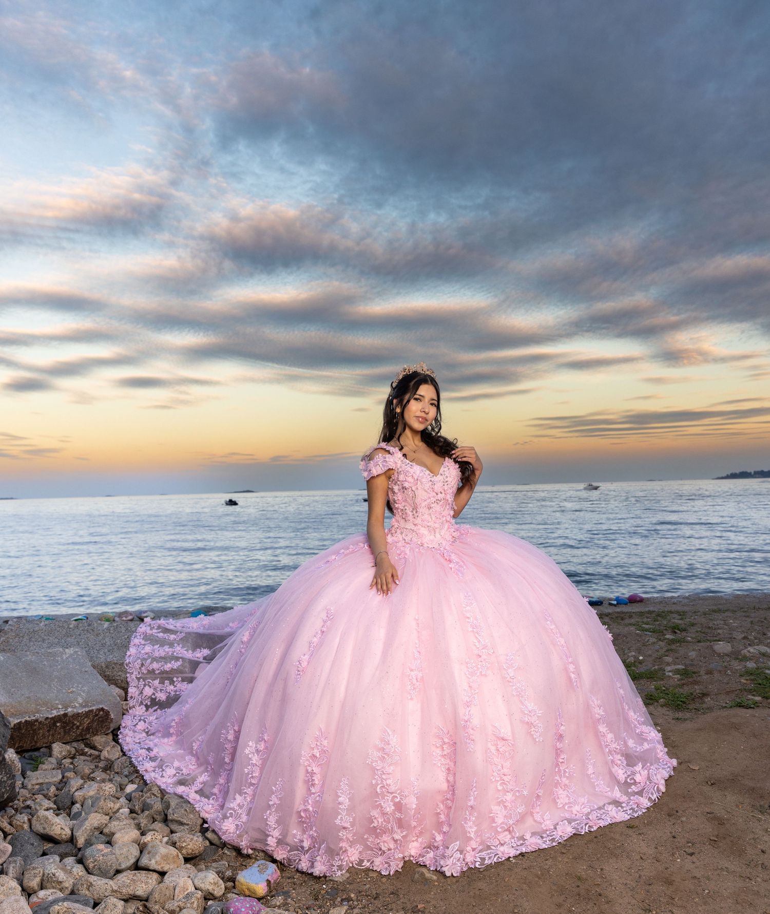 Pink ball gown with floral details photographed against dramatic sunset sky and ocean backdrop.