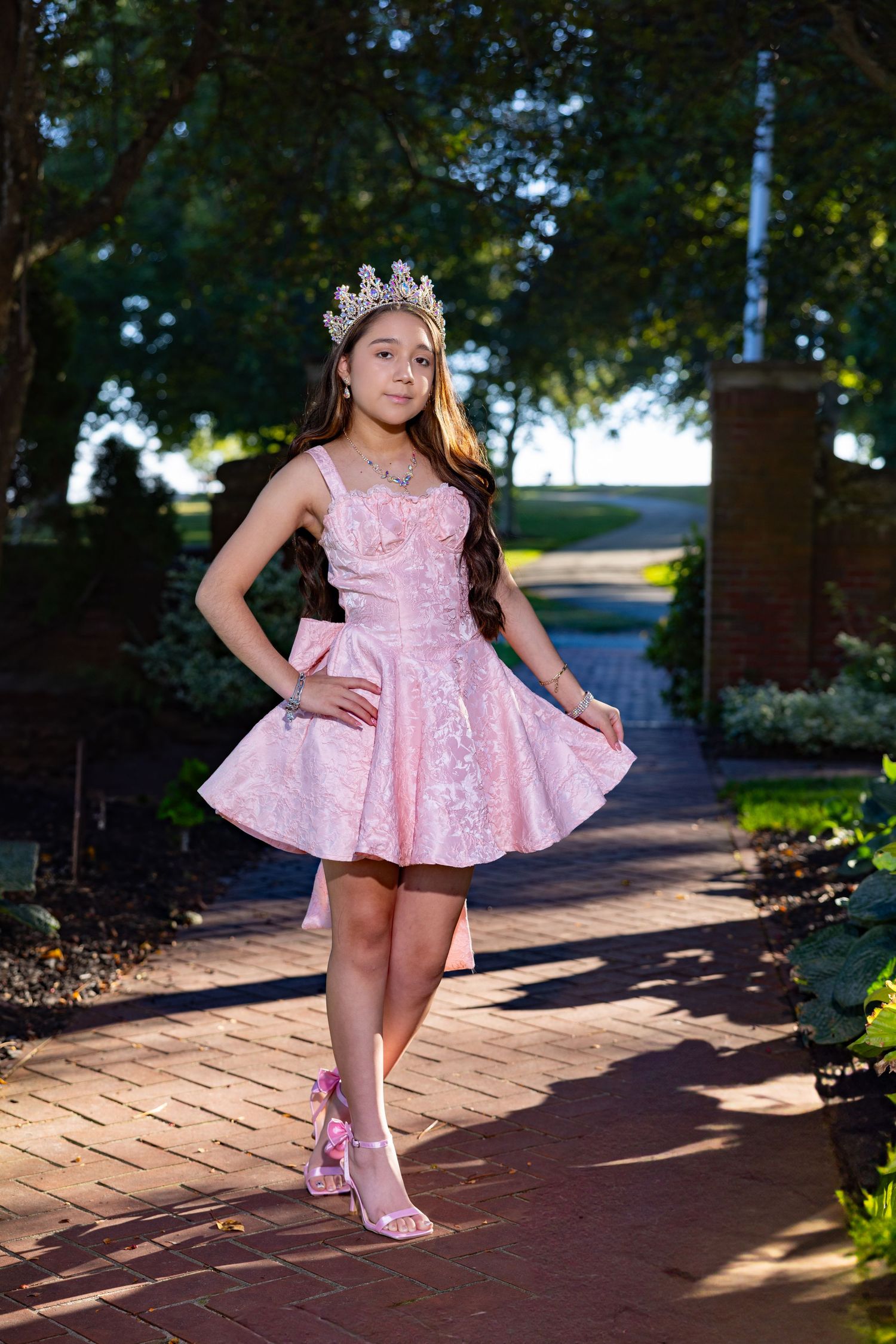 A young dancer in a pink sparkly dress and tiara poses gracefully on a brick pathway in a garden setting.