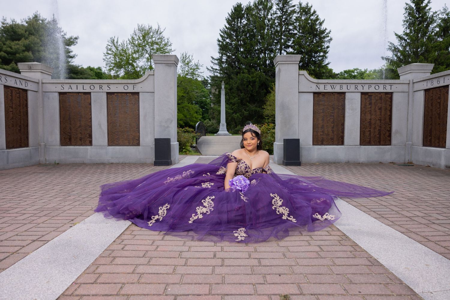 Young woman in elegant purple quinceañera ball gown posing at an outdoor memorial monument surrounded by lush greenery.