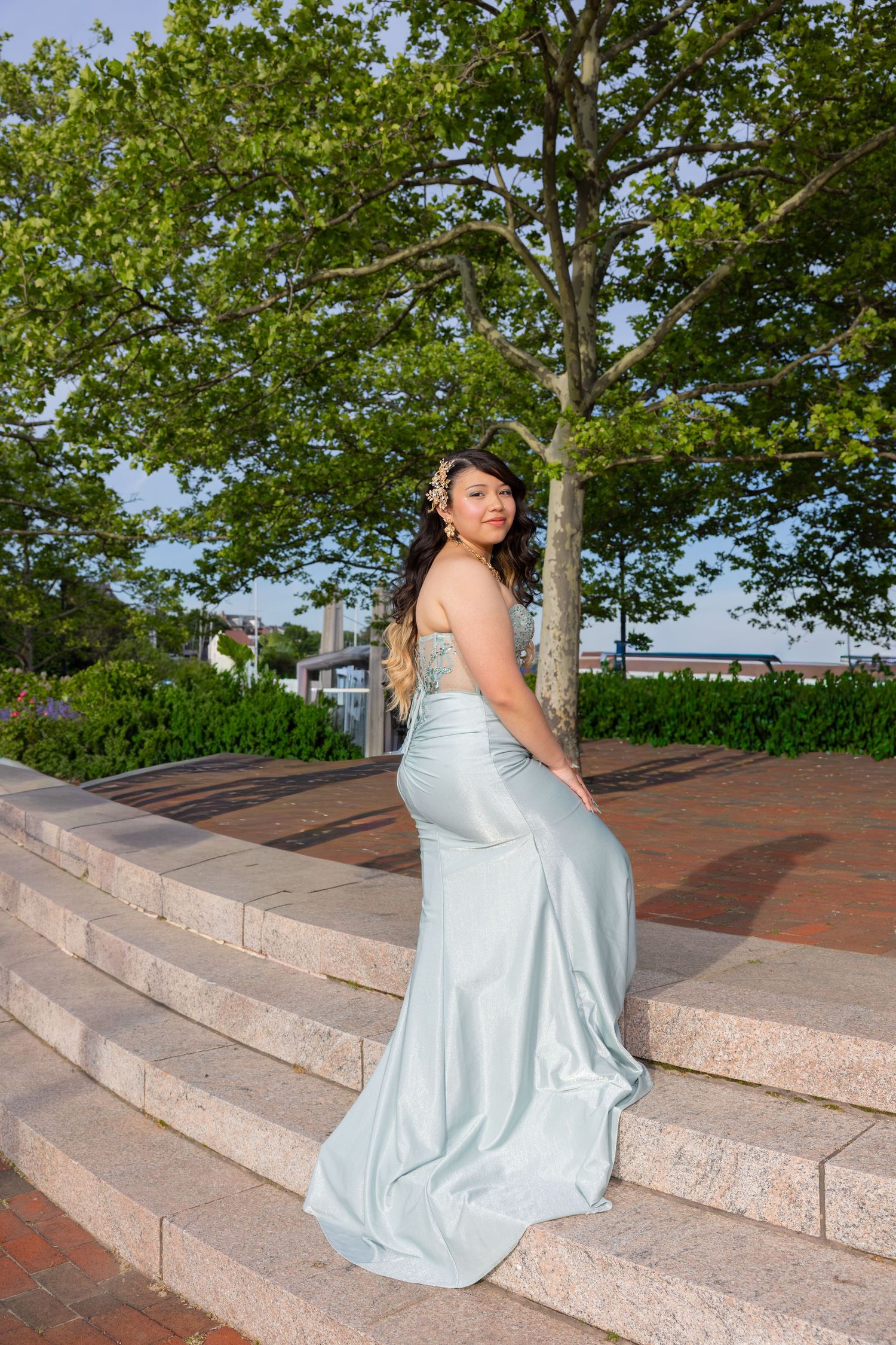 Young woman in elegant light blue satin prom gown sitting on stone steps outdoors with lush green trees behind her.