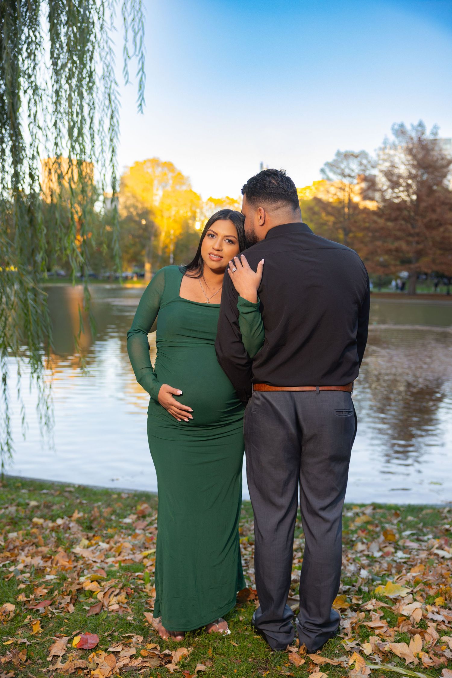 A couple in autumn maternity photos poses together near a lake with willow trees and fallen leaves in golden light.