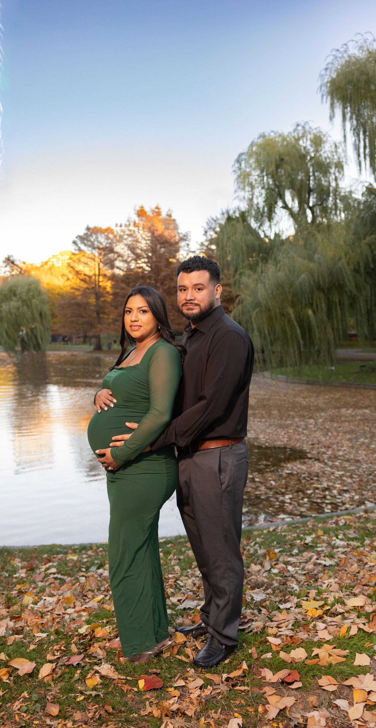 A couple poses for maternity photos by a lake in autumn with fallen leaves scattered on the ground.