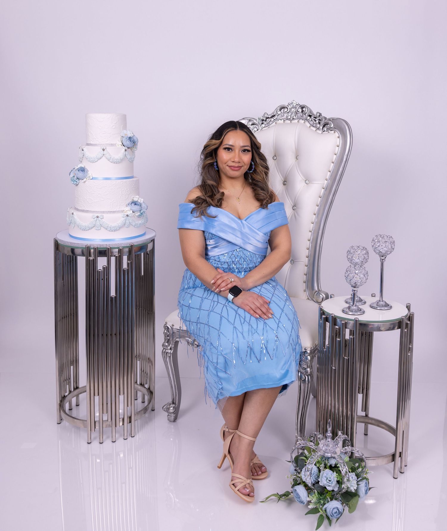 A person in a light blue ruched dress poses elegantly on silver modern furniture against a pale purple backdrop.