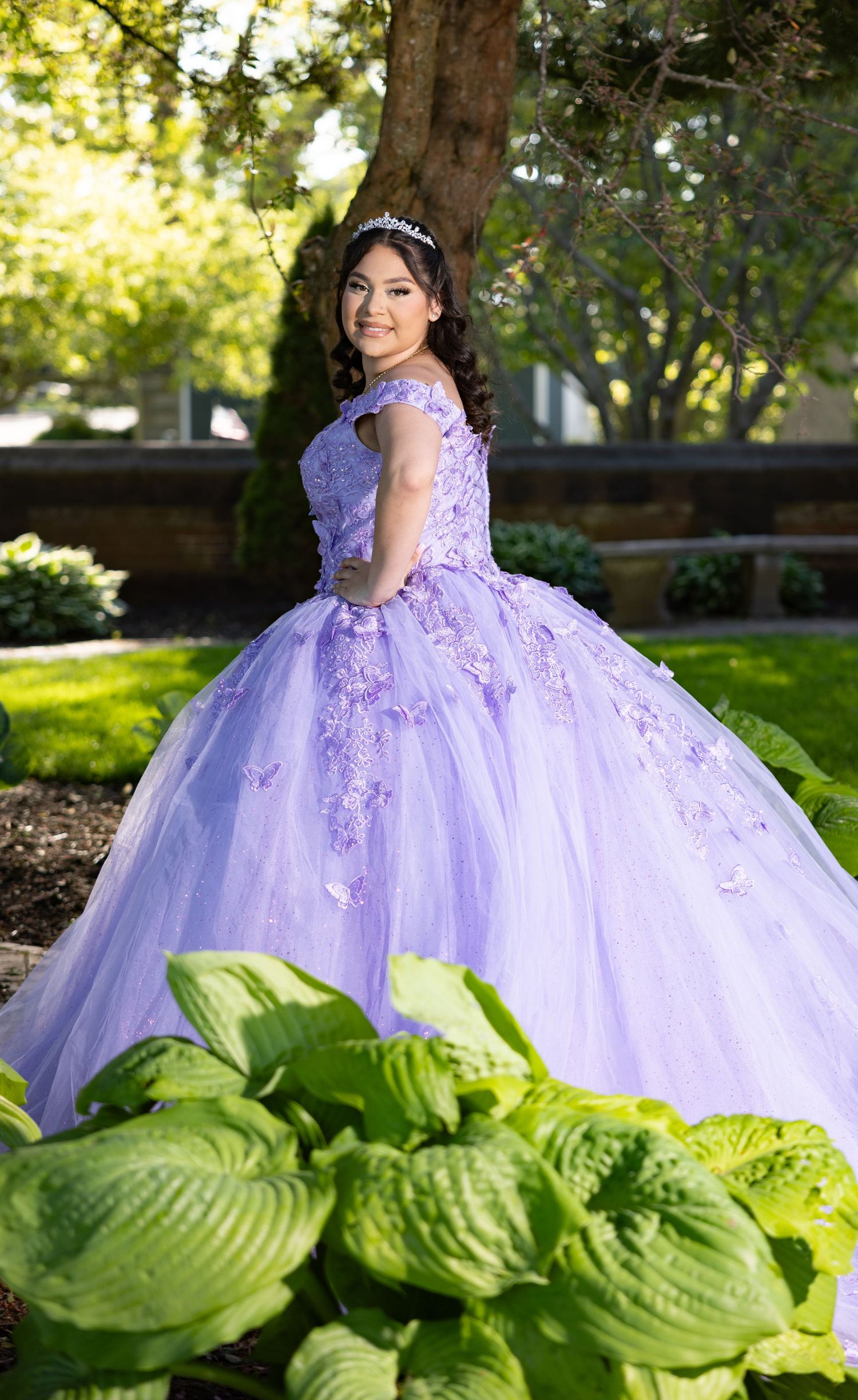 A lavender quinceañera dress with floral appliques photographed in a garden setting with lush green hostas in the foreground.
