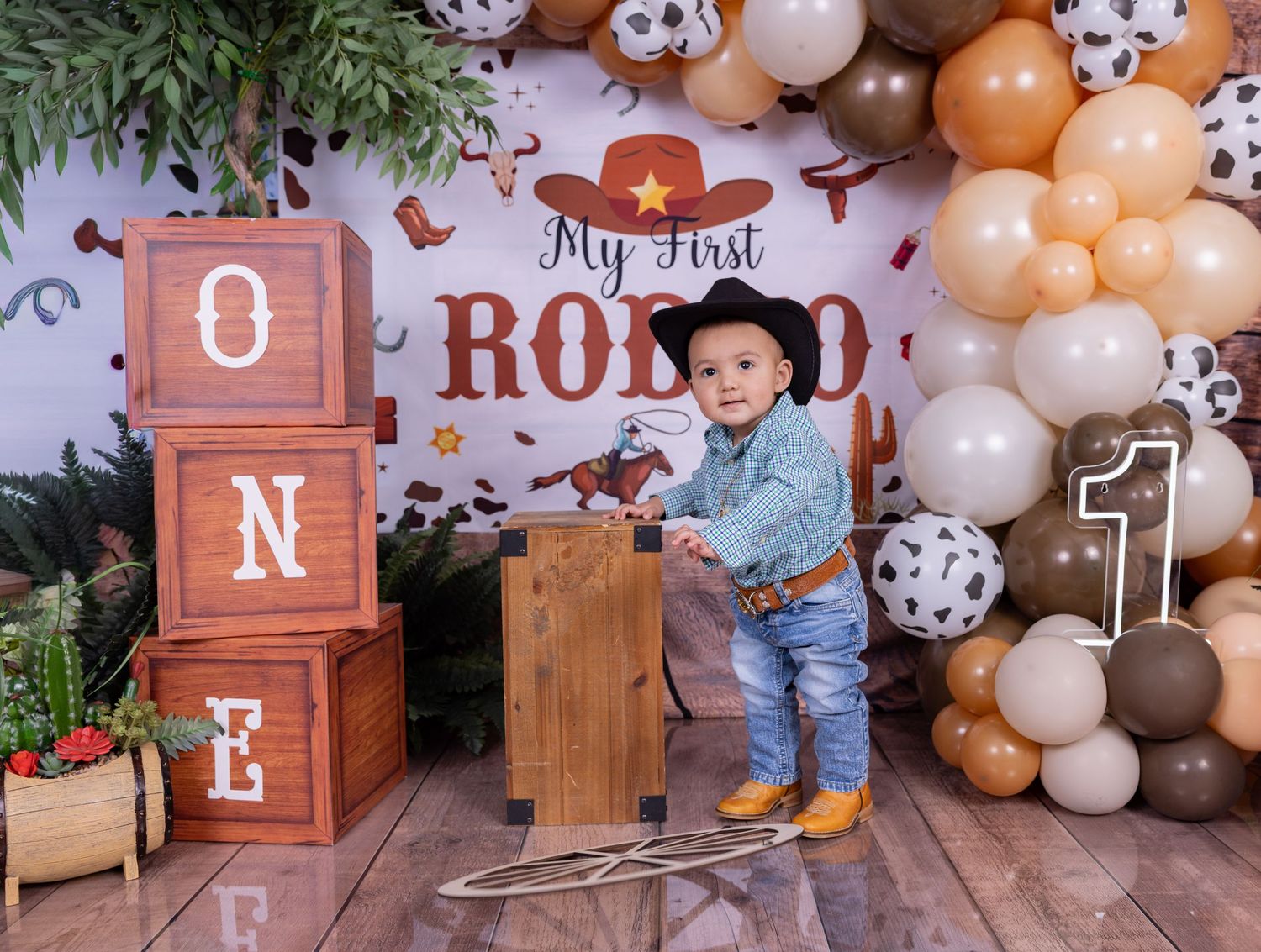 Balloon arch and wooden decorations create a rustic first birthday party setup with brown and white color scheme.