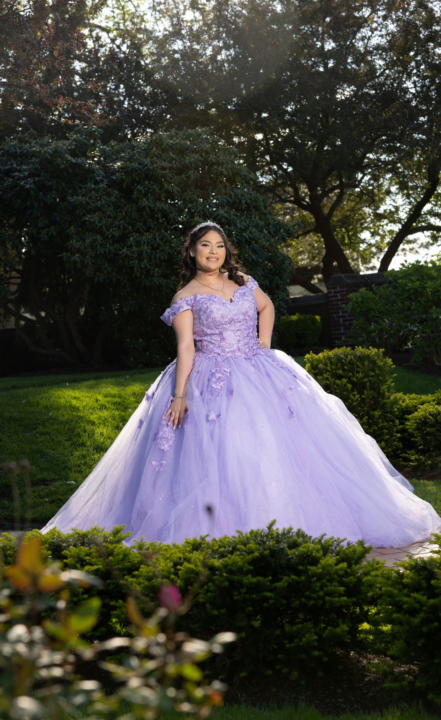 A stunning purple ball gown with delicate lace details captured in an outdoor garden setting with natural sunlight filtering through.