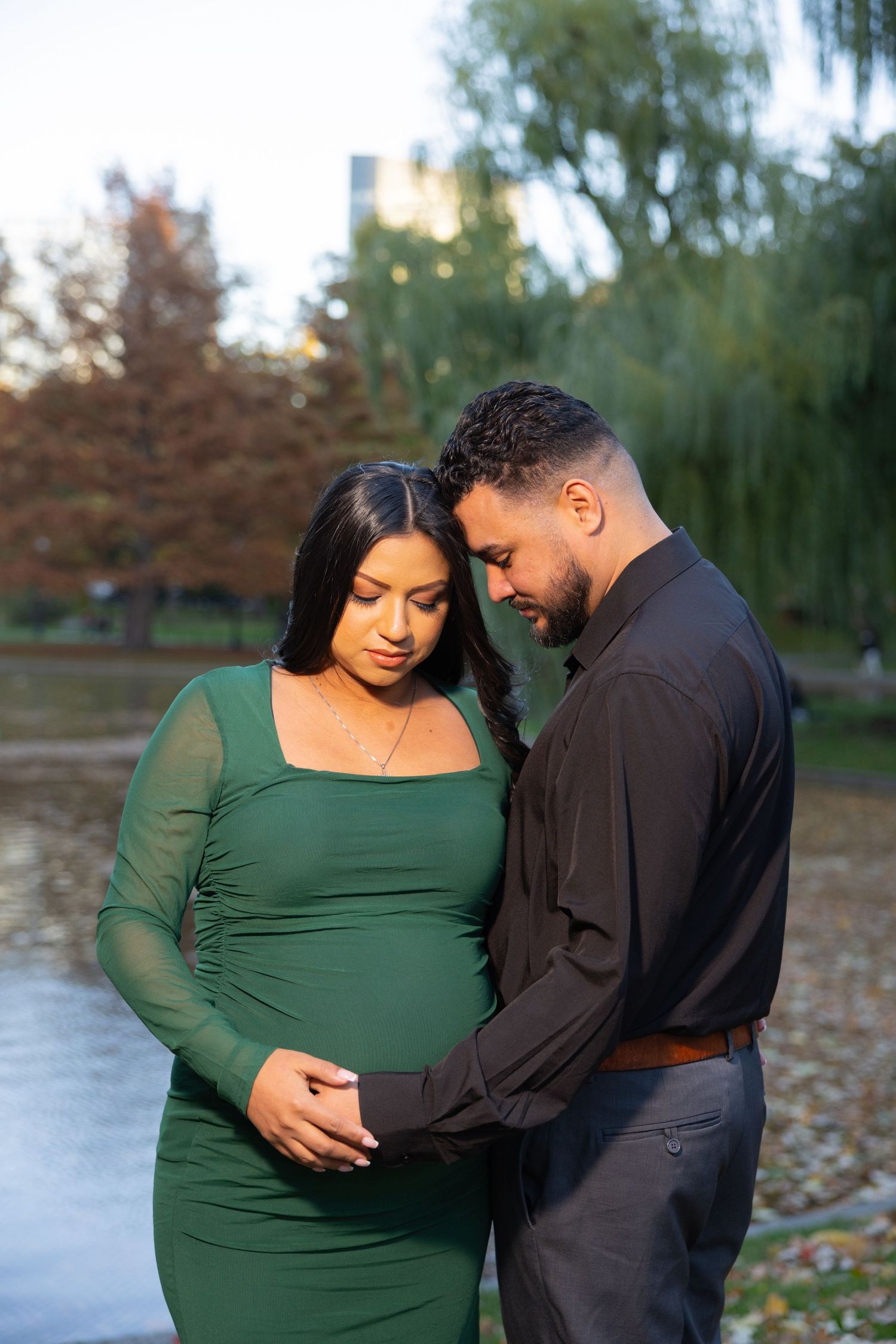 Couple in black and green outfits share an intimate moment during maternity photoshoot in autumn park setting.
