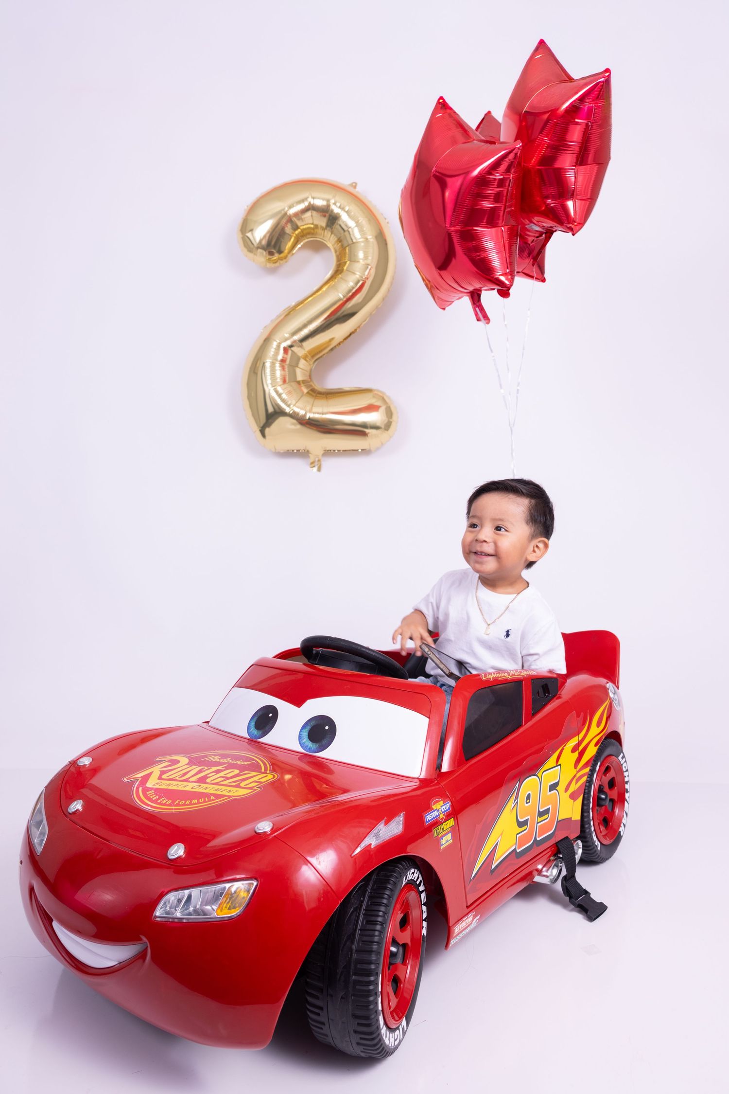 Toddler boy sitting in a red Lightning McQueen ride-on car with a gold number 2 balloon for his 2nd birthday.