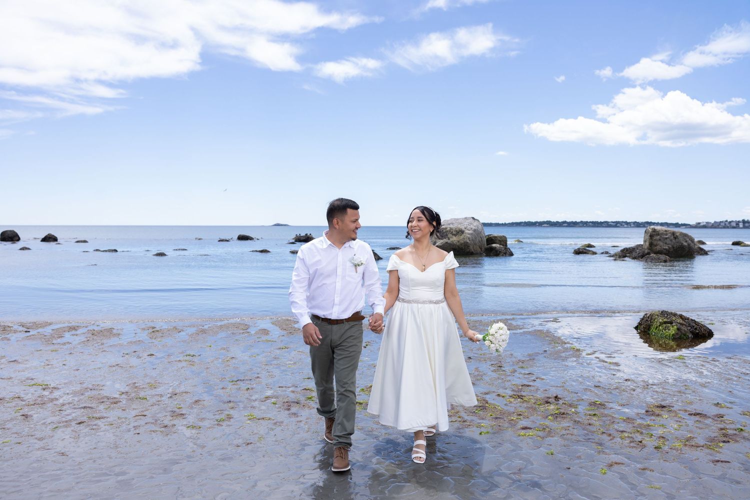 Bride and groom walk hand-in-hand along a scenic beach shoreline on their wedding day.