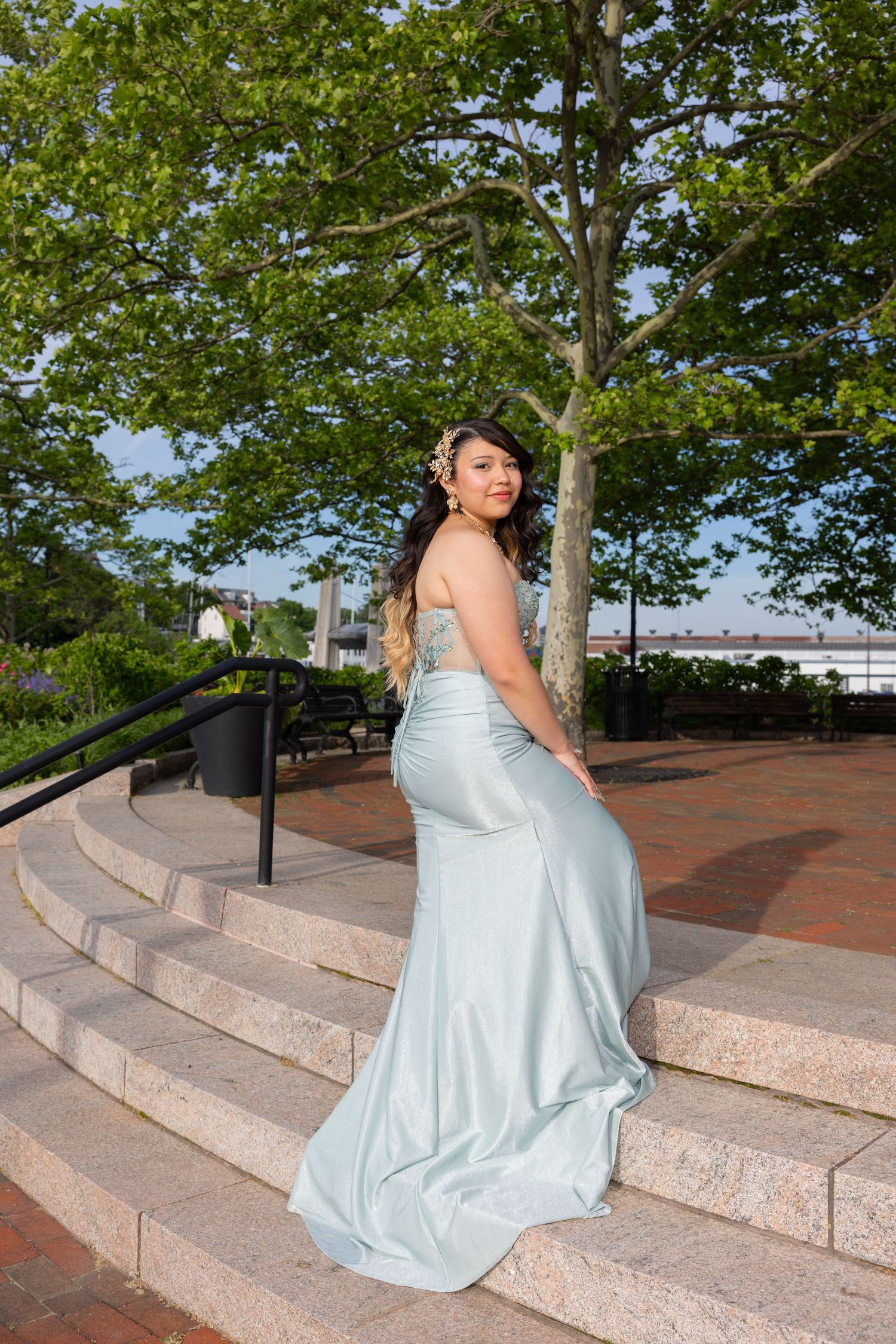 Young woman in elegant light blue satin mermaid gown poses on stone steps outdoors among green trees.