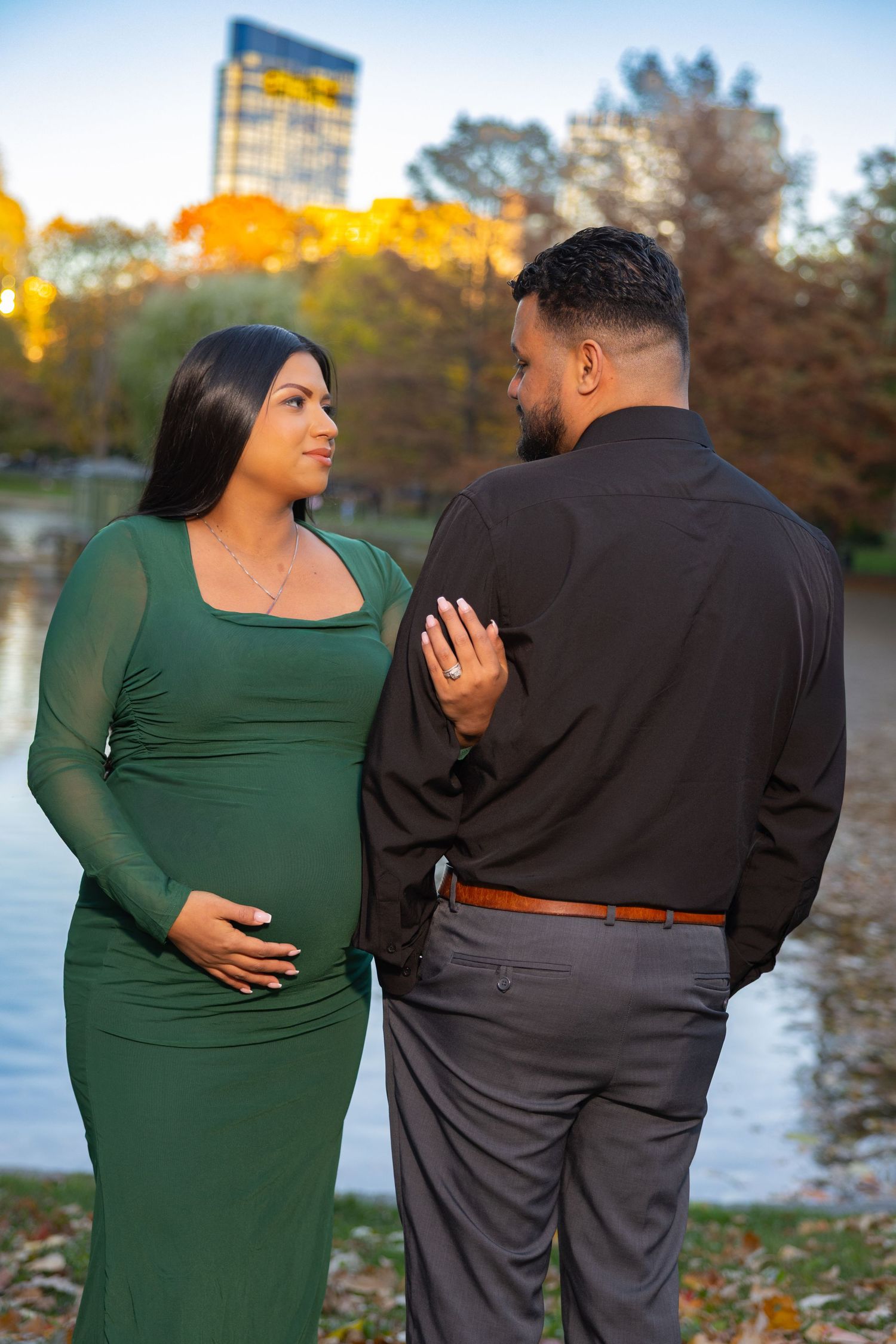 A couple in formal attire poses lovingly by a lake during autumn, with the person in green touching their stomach.