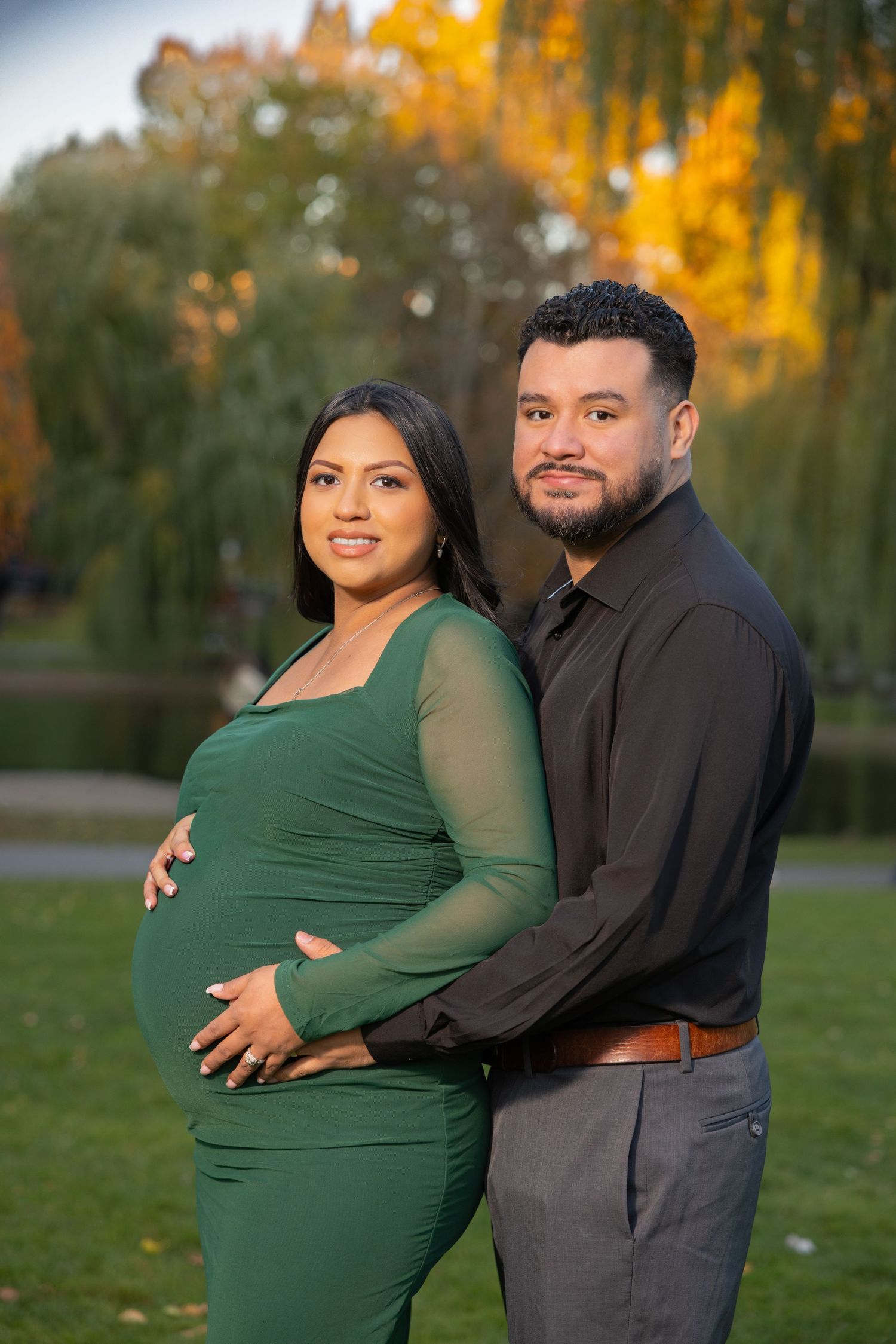 Expectant couple poses outdoors in autumn setting with golden foliage as woman in green dress cradles baby bump.